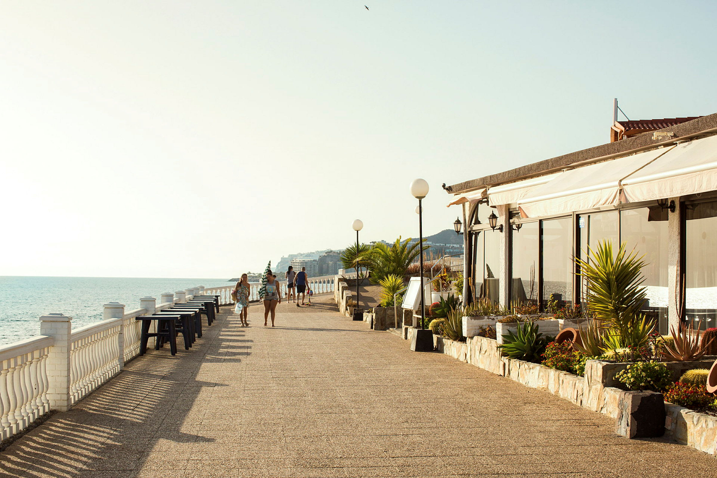 Strandpromenade med havudsigt, borde langs rækværket og folk der går tur. (AI-genereret tekst)