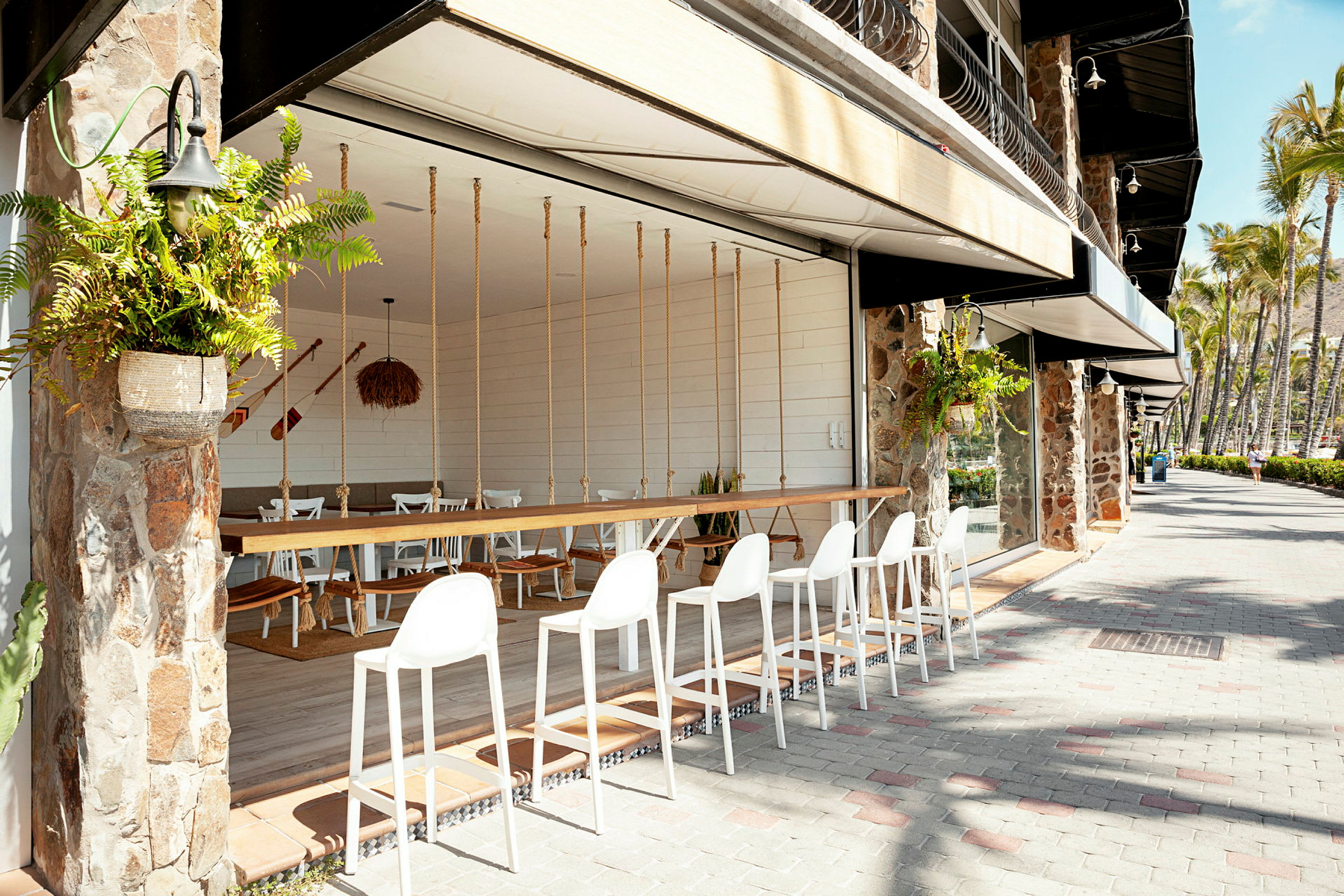 Udendørsområde med hvide barstole ved en bænk, med plantepynt og en restaurant med siddepladser i umiddelbar nærhed. (AI-genereret tekst)