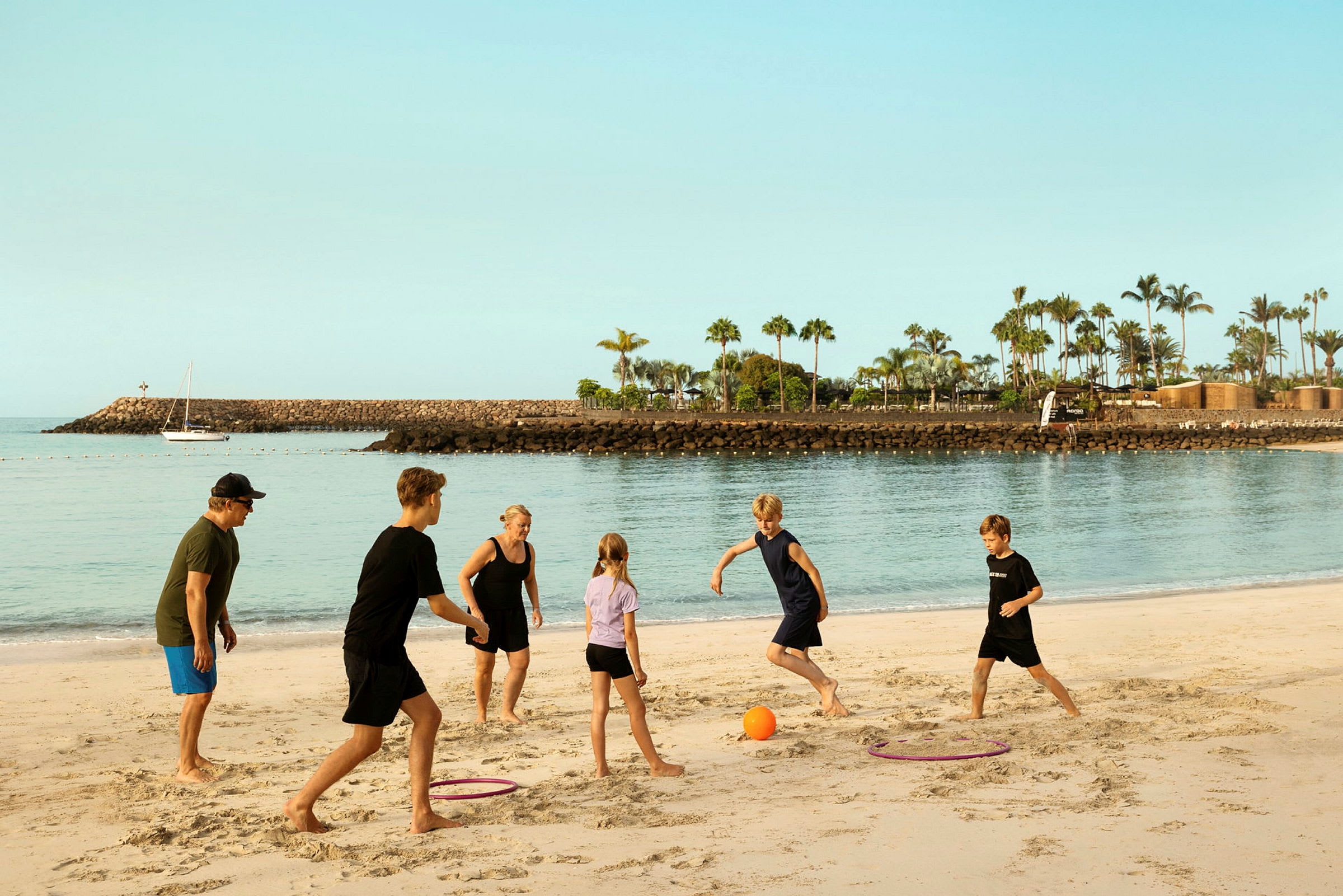 En gruppe personer leger med en bold på en sandstrand med en havn og palmer i baggrunden. (AI-genereret tekst)
