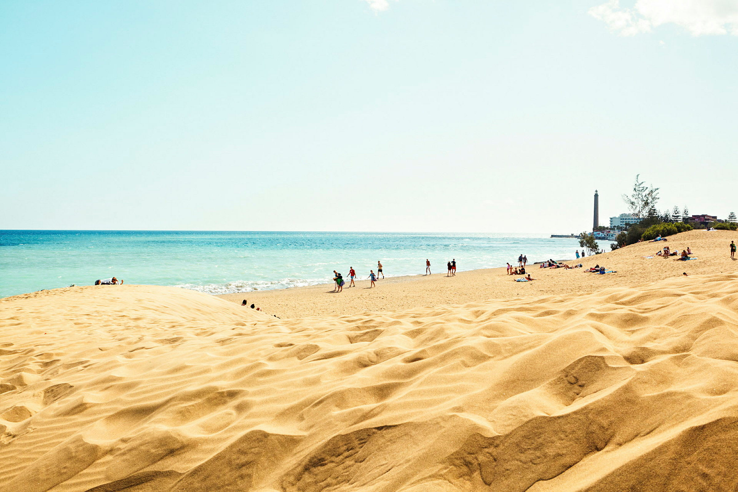 Strand med sandklitter og mennesker tæt ved havet, med et fyrtårn til højre. (AI-genereret tekst)