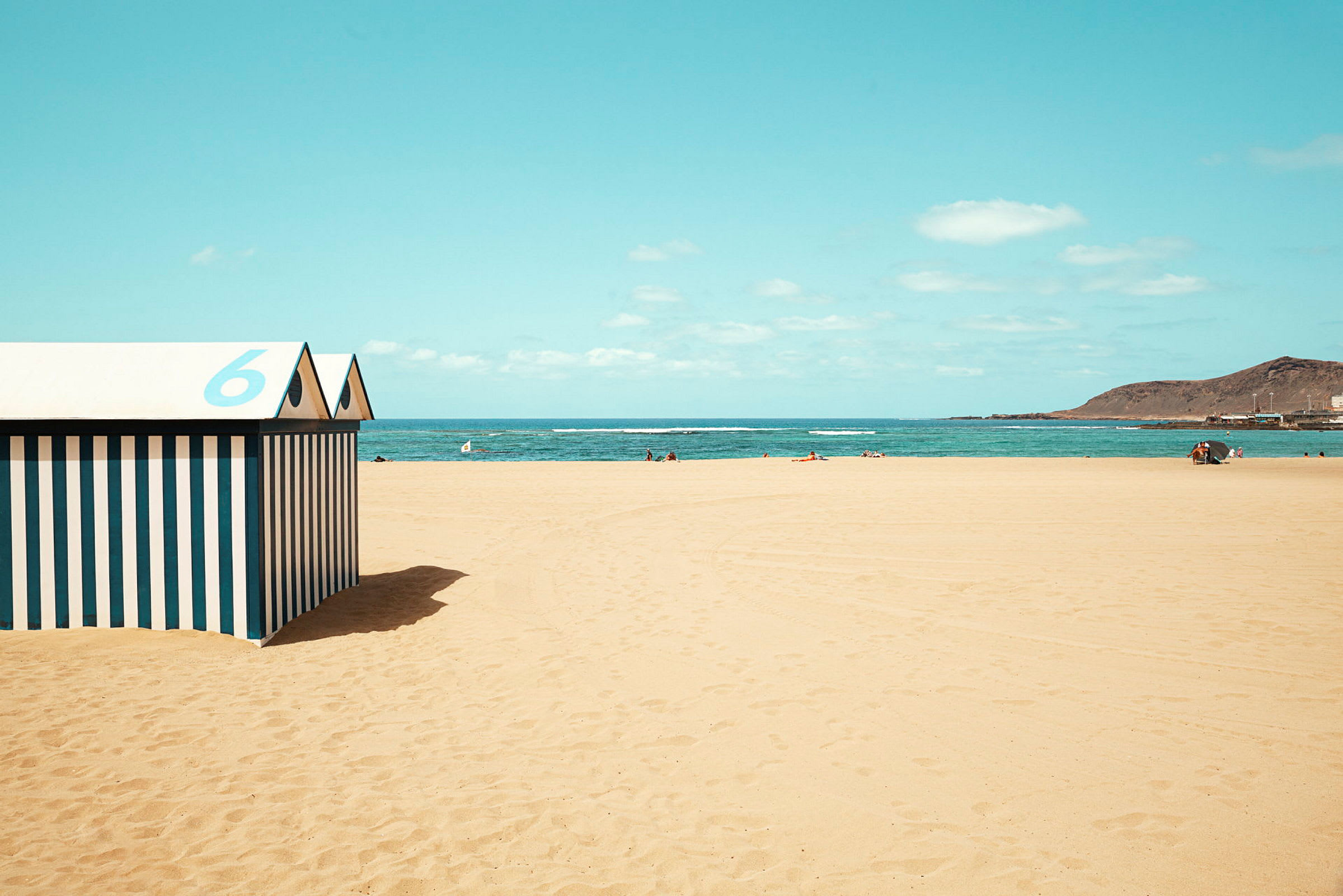 Strand med gul sand, blåt hav og himmel samt et stribet strandhus med nummer 6 i forgrunden. (AI-genereret tekst)