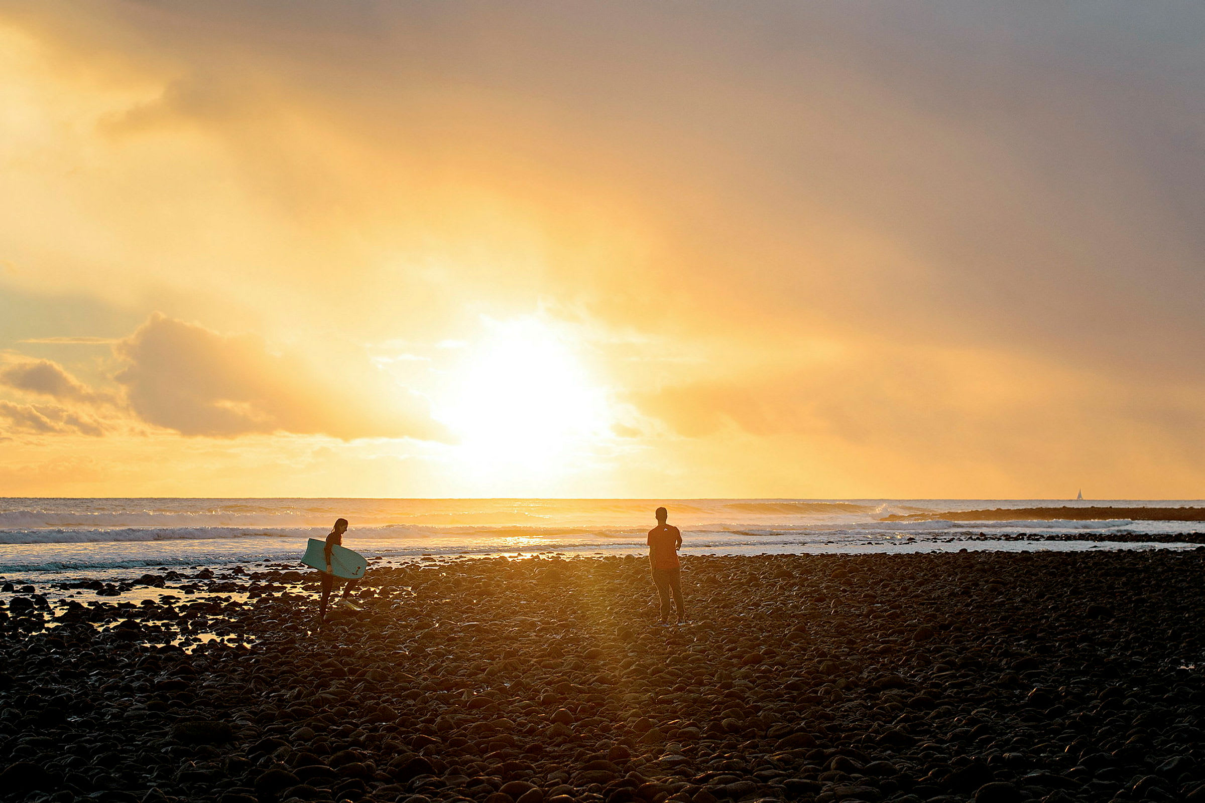 En solnedgang over havet med to personer på en stenstrand, den ene med et surfbræt. (AI-genereret tekst)