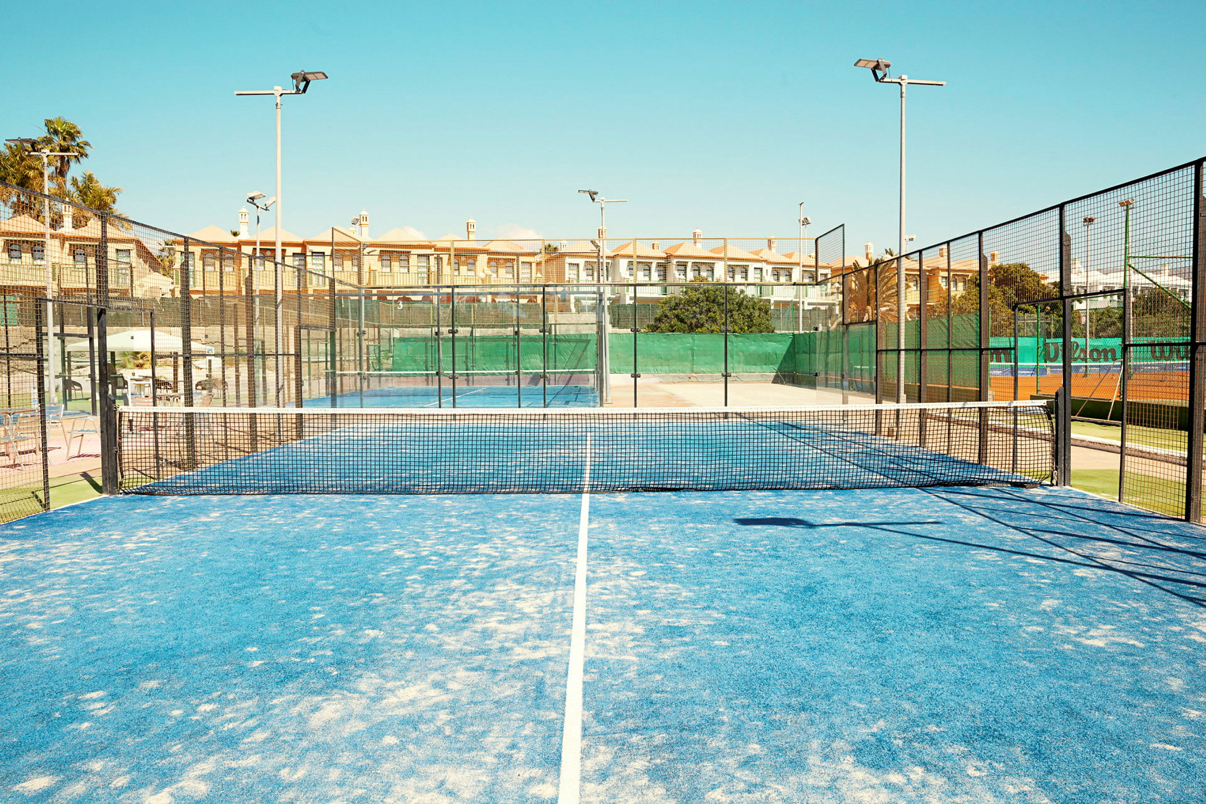En padelbane med blåt underlag og net i midten, omgivet af hegn og bygninger i baggrunden. (AI-genereret tekst)