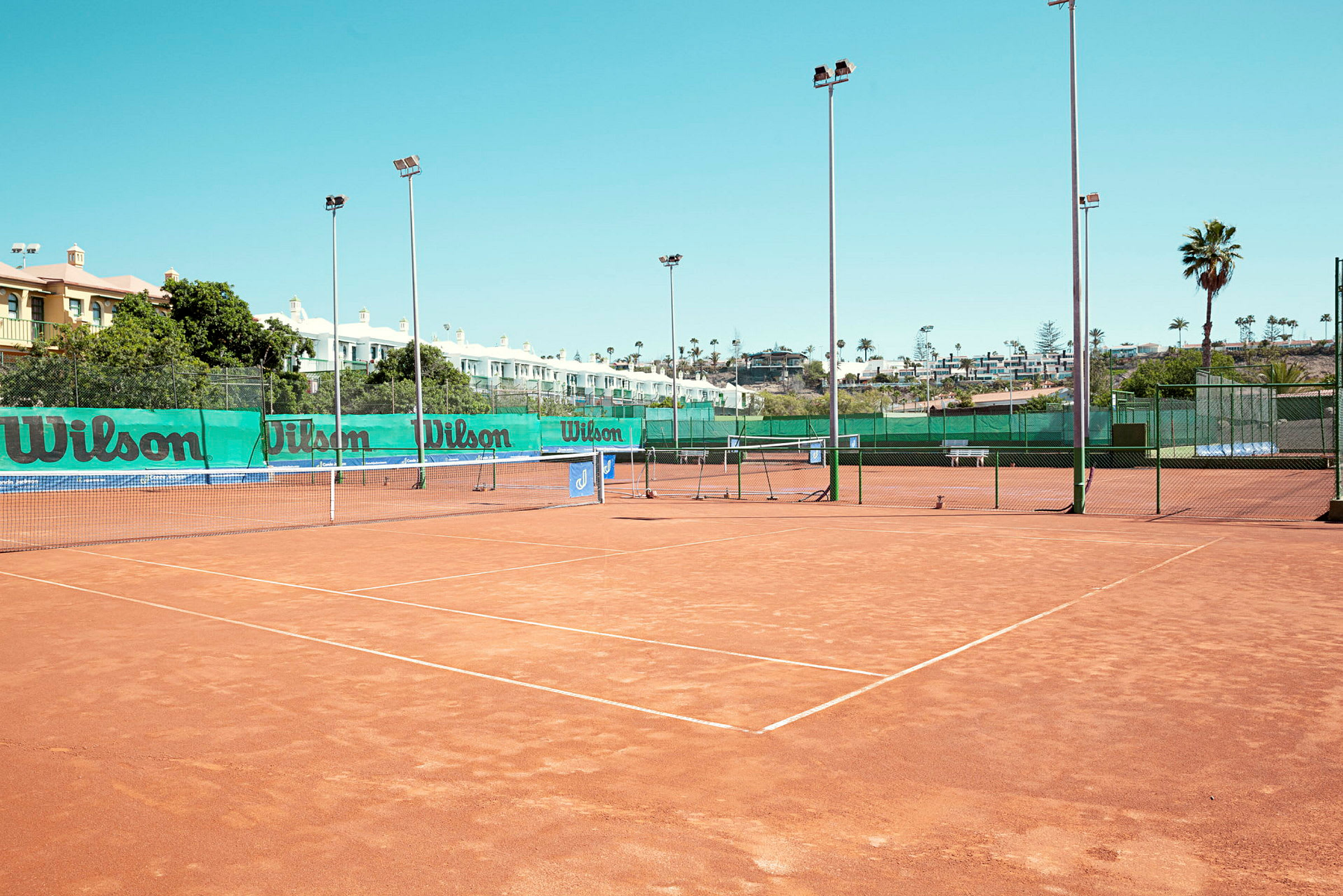 Tennisbaner med grusunderlag, omgivet af net og lysmaster. (AI-genereret tekst)
