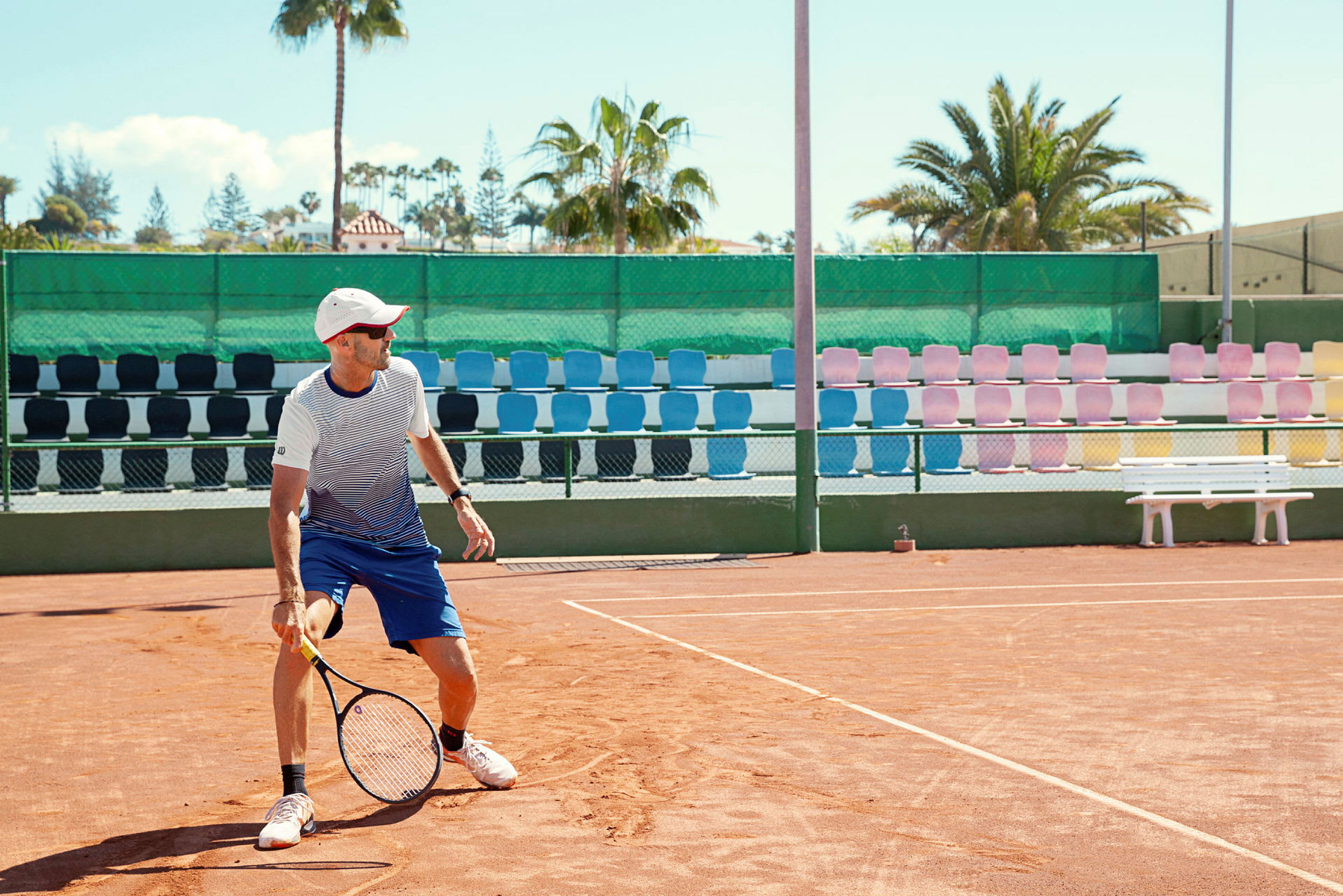 En tennisbane med en spiller, der holder en ketcher, omgivet af farverige sæder og palmer i baggrunden. (AI-genereret tekst)