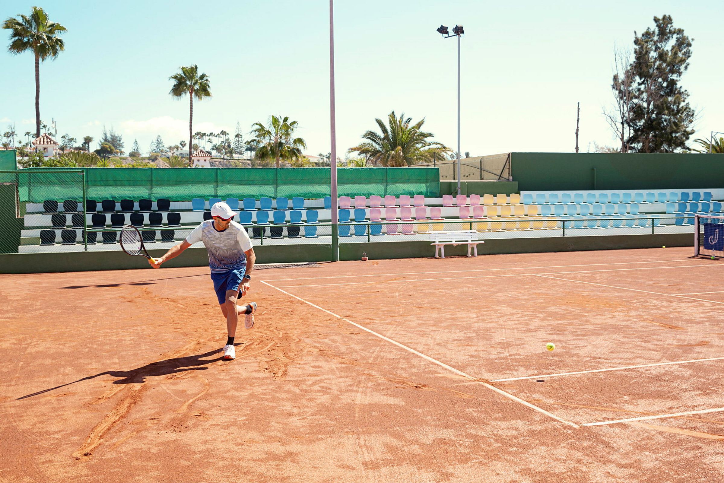 En person spiller tennis på en udendørs grusbane med farverige tribunestole og palmer i baggrunden. (AI-genereret tekst)