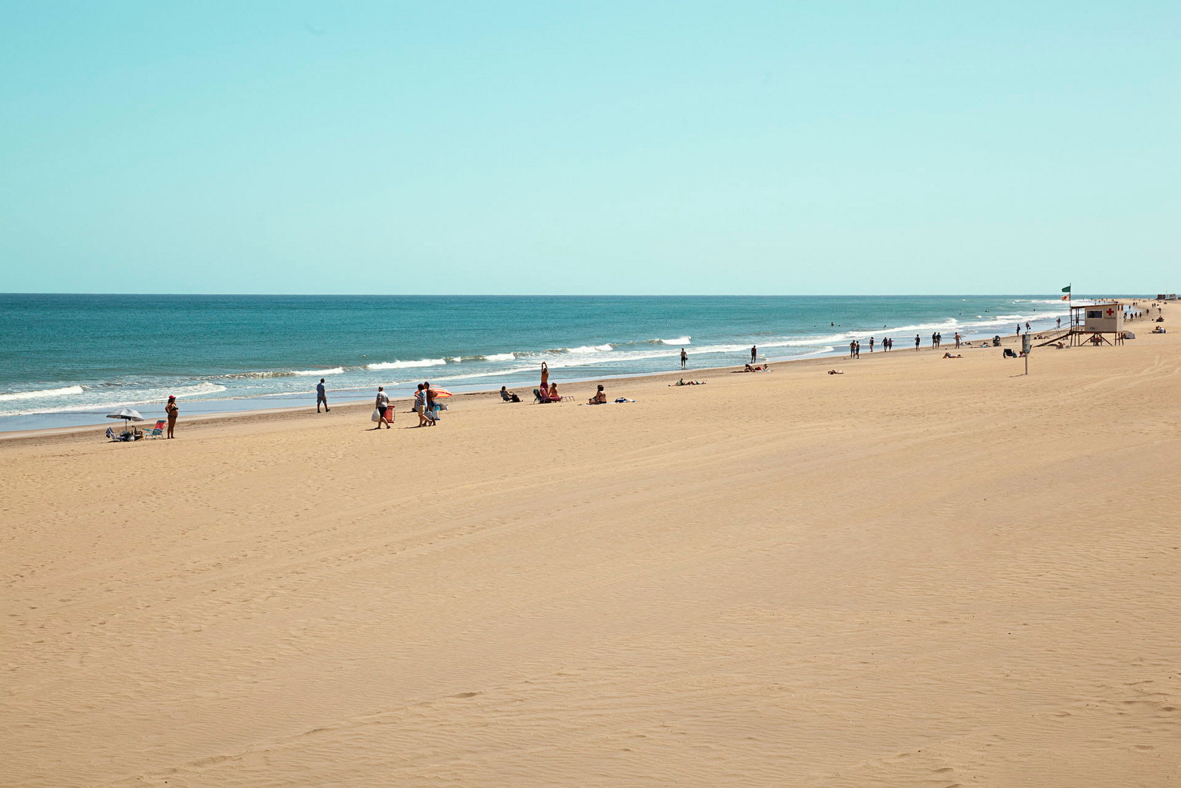 En lang sandstrand med mennesker, der nyder solen og havet, samt en livreddelsesstation. (AI-genereret tekst)