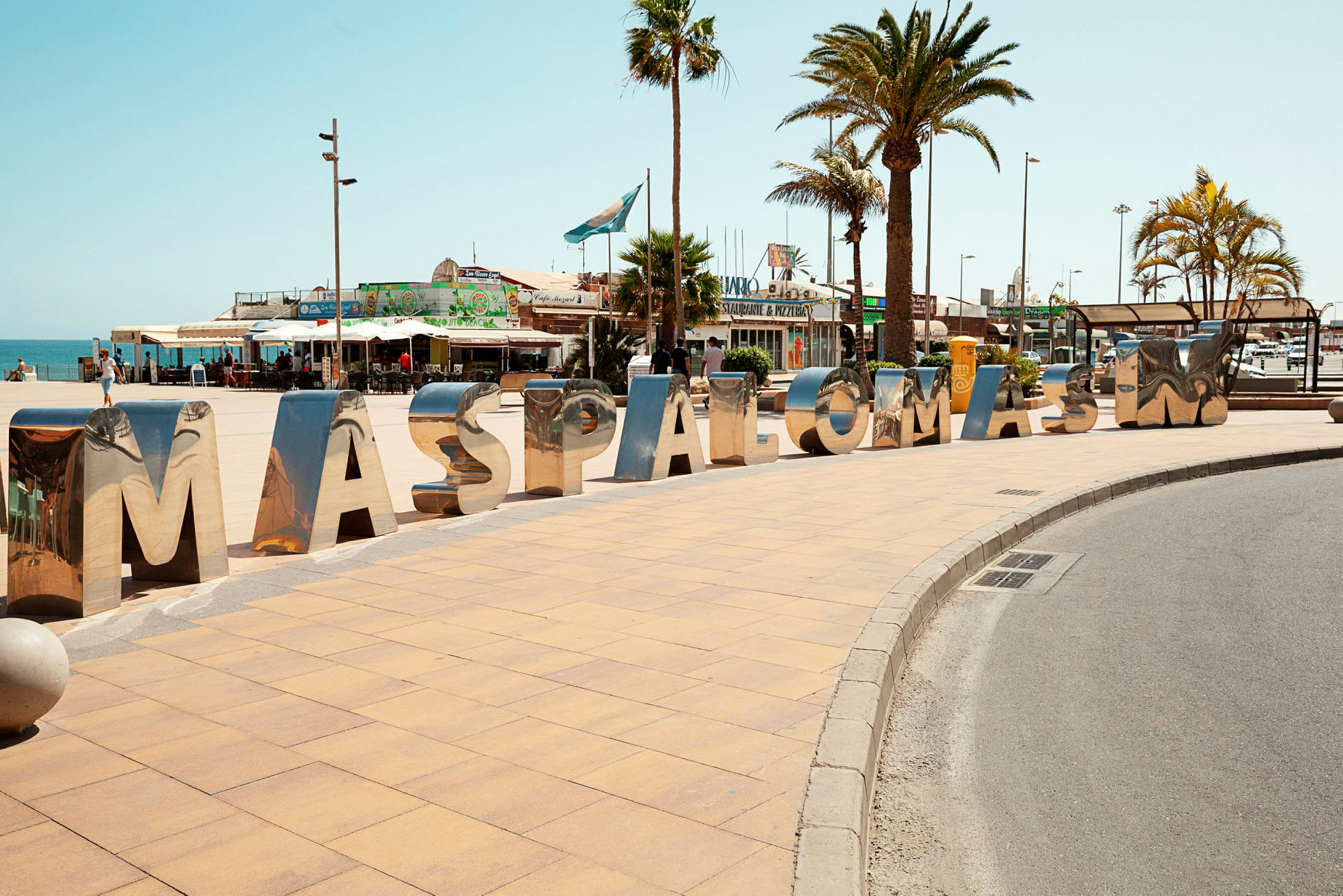 Store, blanke bogstaver udgør ordet "MASPALOMAS" på en fortov ved en plads med palmer. (AI-genereret tekst)