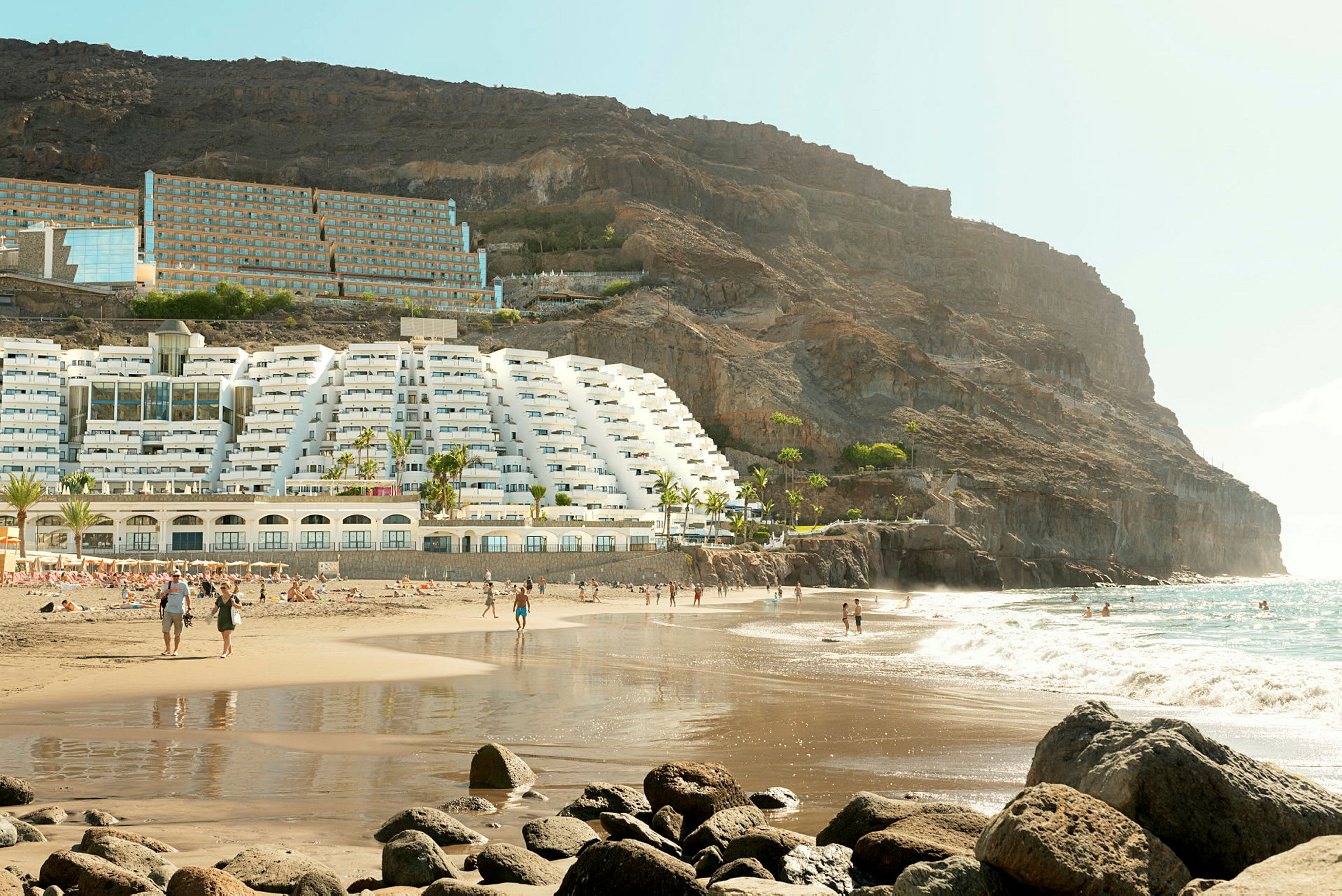 Strand med hotelbygning ved en skråning og mennesker, der bevæger sig langs sanden og vandet. (AI-genereret tekst)