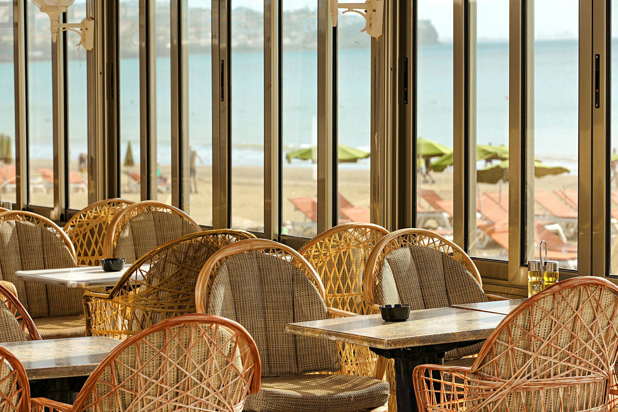 Udsigt fra en restaurant med fletstole og borde samt panoramavinduer med udsigt over stranden og havet. (AI-genereret tekst)