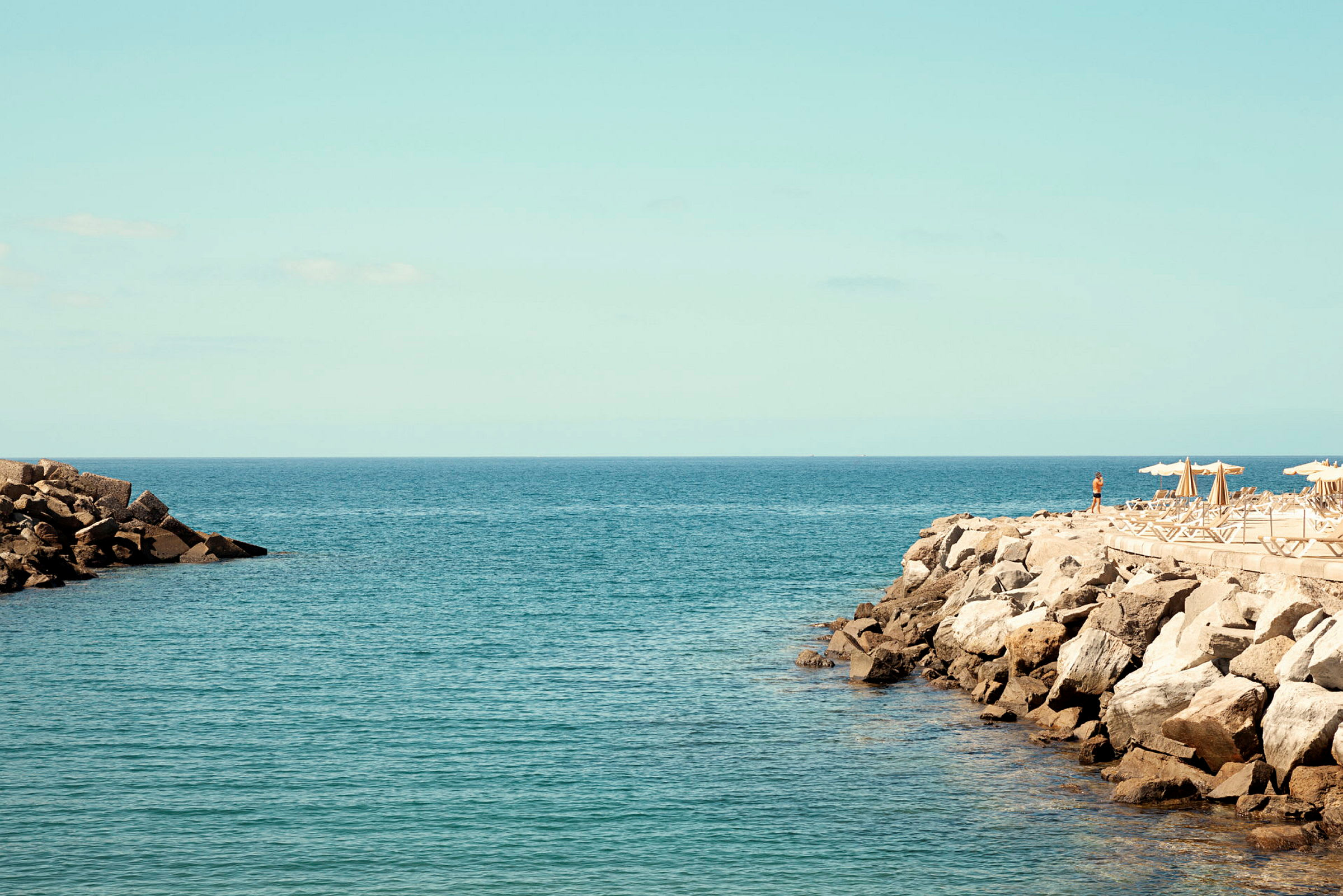 Udsigt over havet med stenet kyst og liggestole under parasoller. (AI-genereret tekst)
