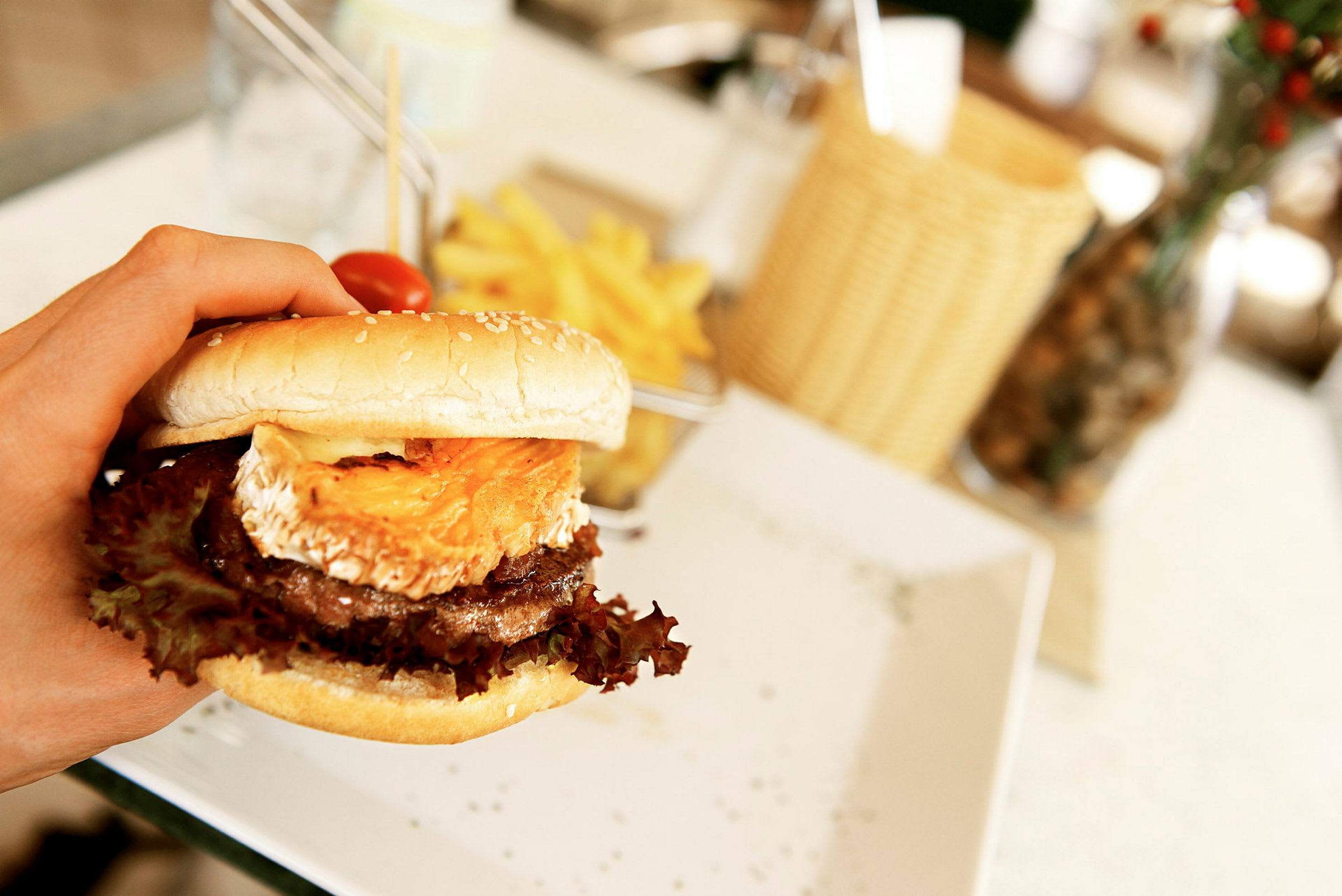 En hamburger med ost og salat holdes i en hånd, med pommes frites på en tallerken i baggrunden. (AI-genereret tekst)