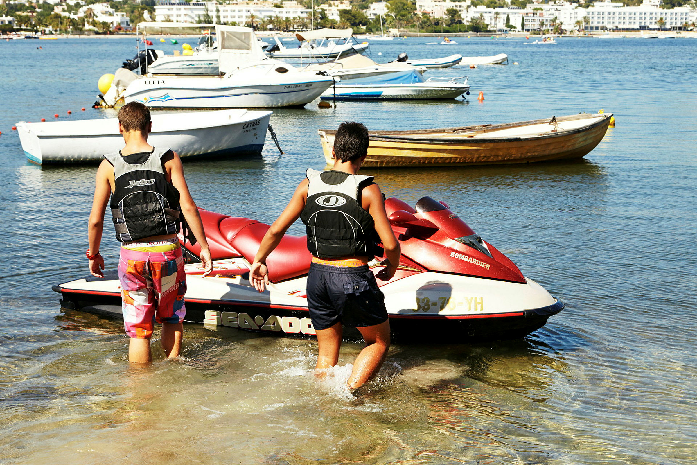 To personer trækker en vandscooter ud i vandet på en strand med flere både fortøjet i baggrunden. (AI-genereret tekst)