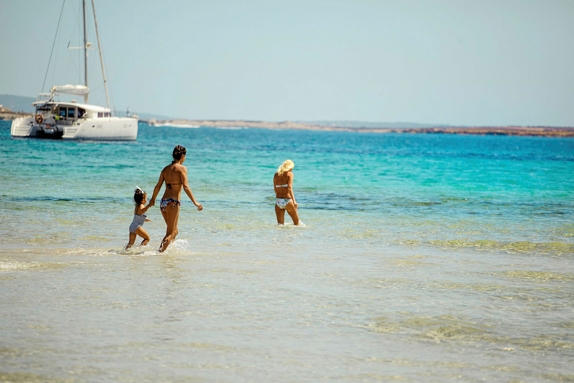 Tre personer går i lavt, klart vand på en strand, med en sejlbåd i baggrunden. (AI-genereret tekst)
