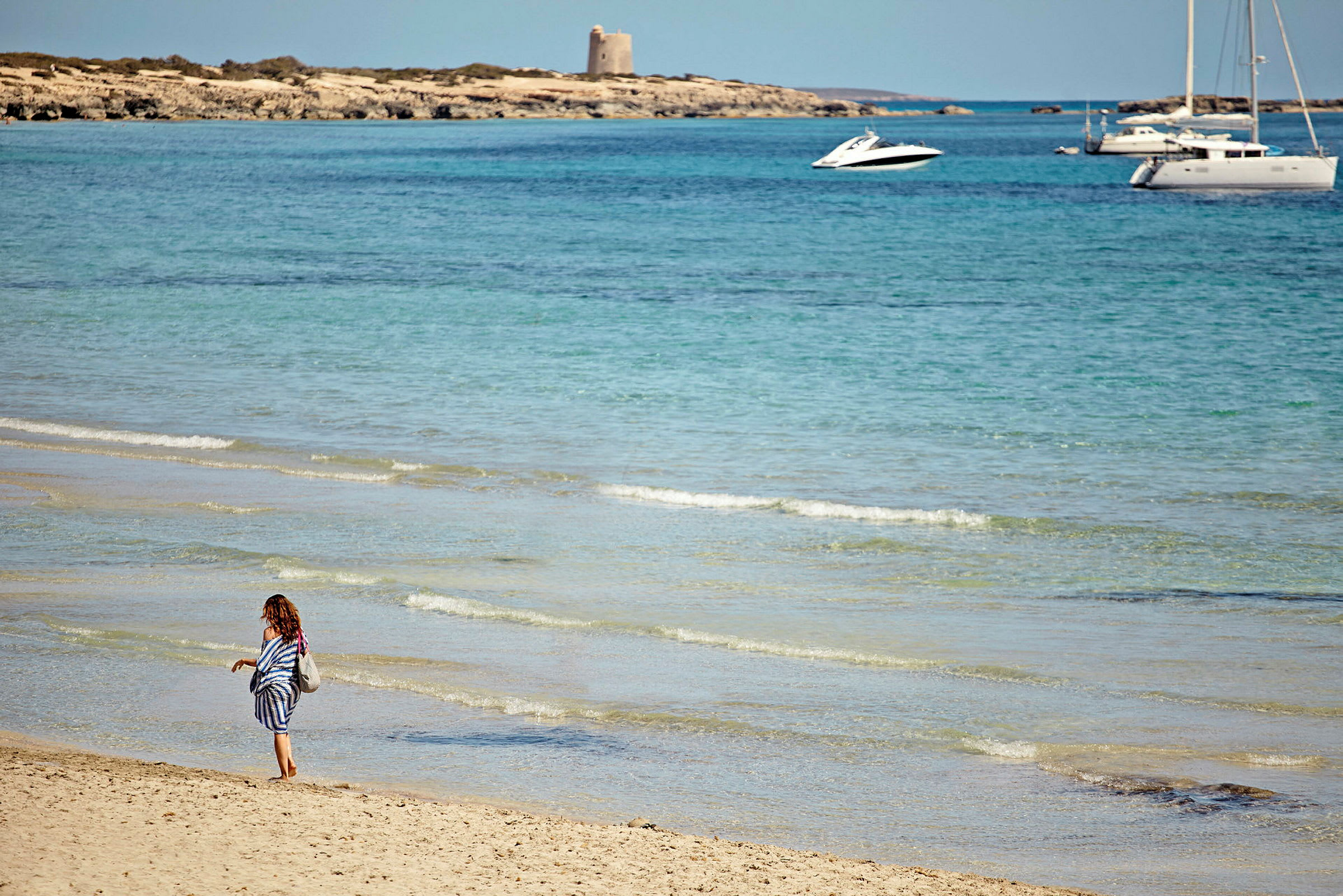 En person går langs en sandstrand ved havet med klare bølger. I baggrunden ses en lille båd og en sejlbåd ude på vandet samt en klippe med en bygning. (AI-genereret tekst)