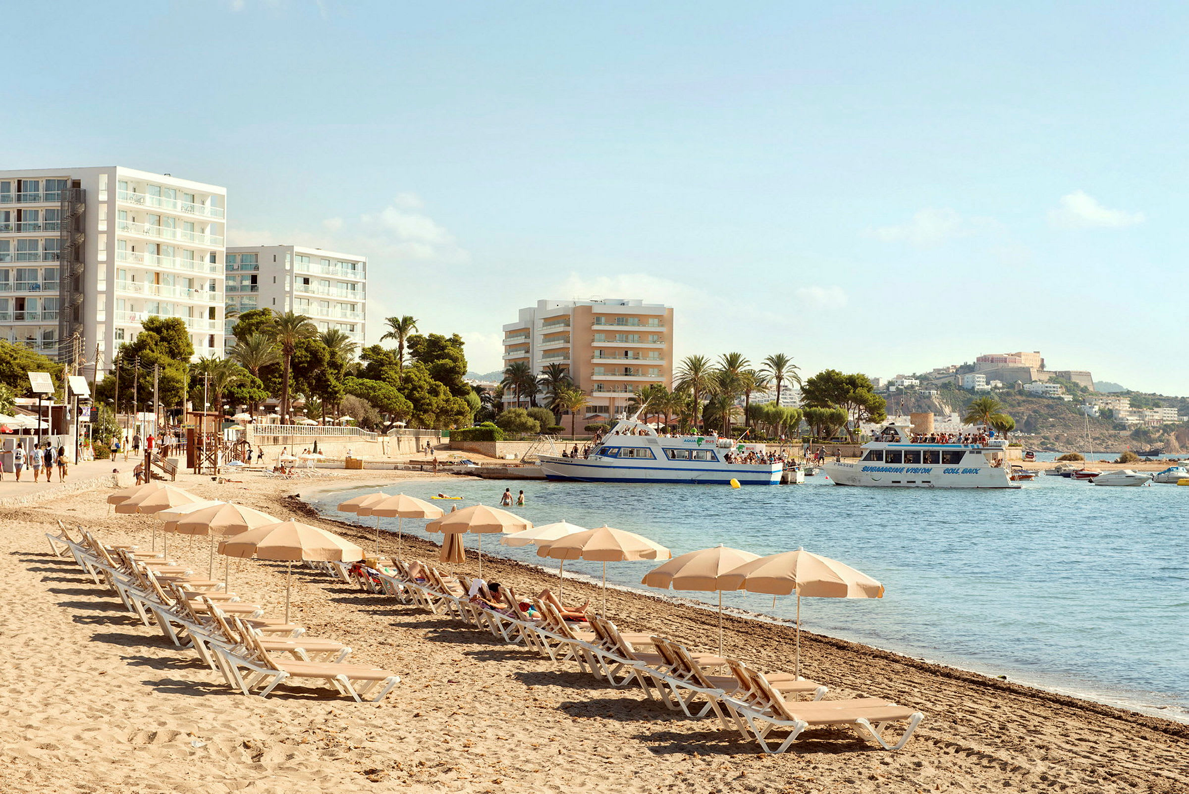 Strand med liggestole og parasoller, hotelbygninger i baggrunden og både på vandet. (AI-genereret tekst)