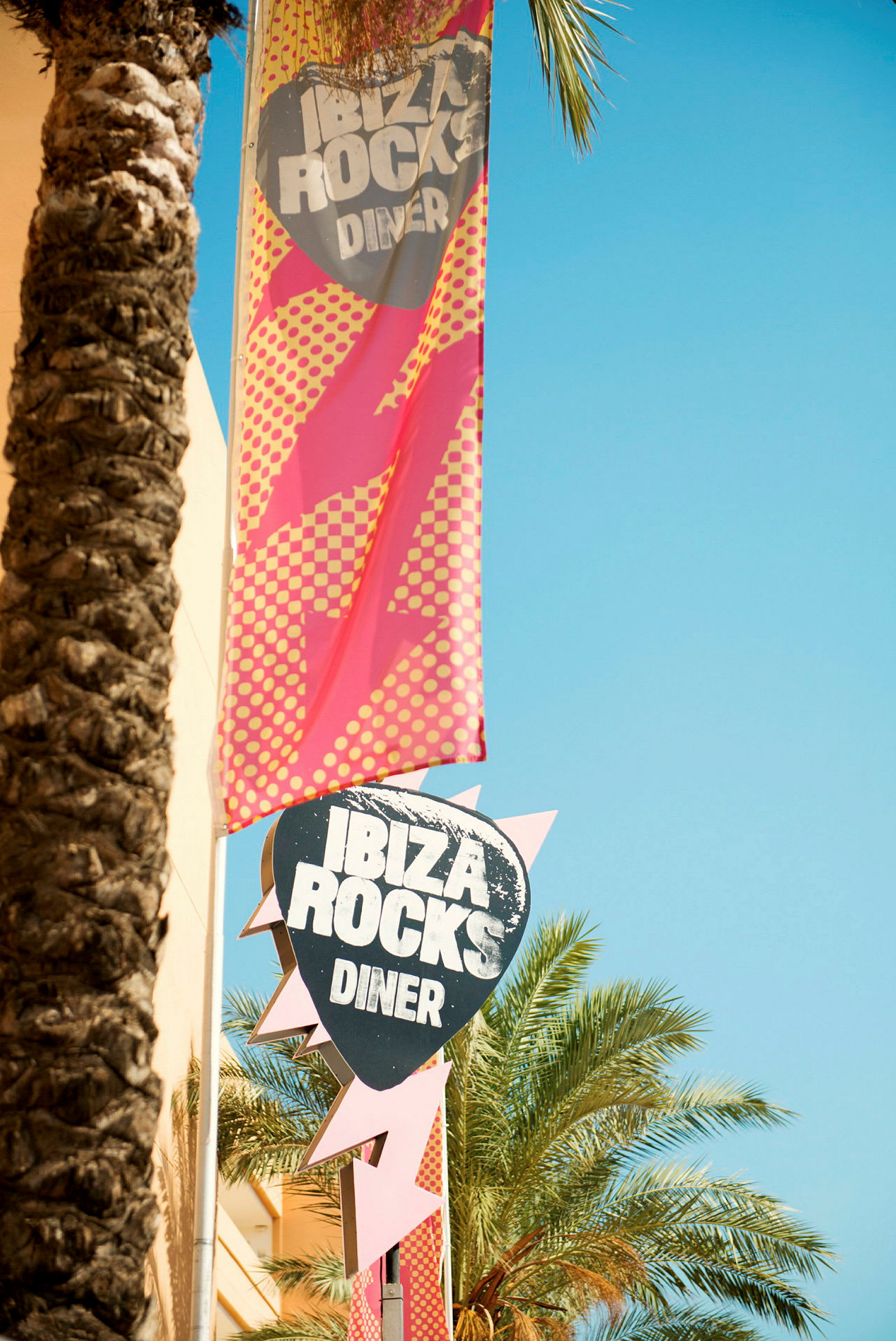 Et skilt og banner for Ibiza Rocks Diner, omgivet af palmer og en klar himmel. (AI-genereret tekst)