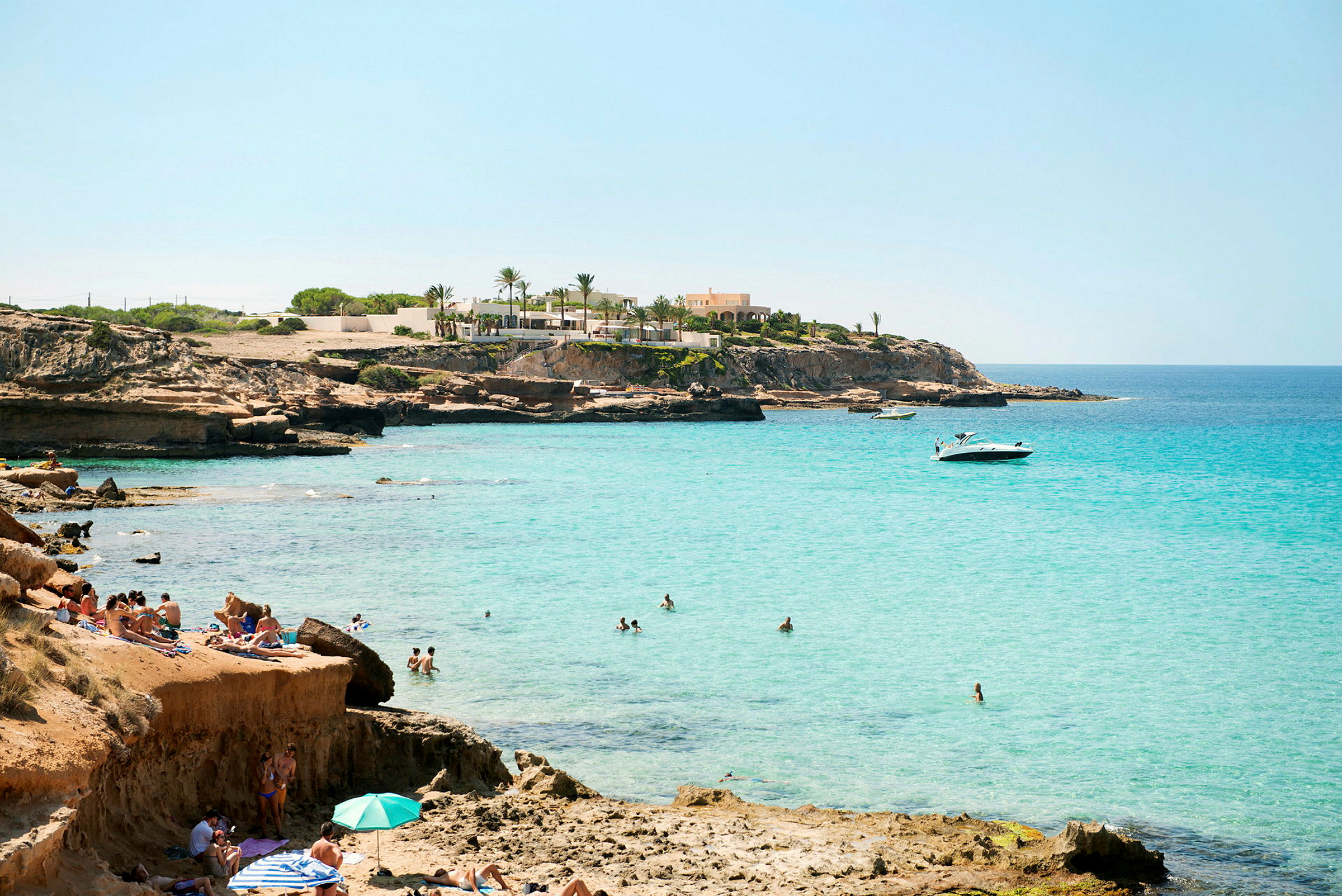 En klippefyldt strand med turkisblåt vand og mennesker, der bader og soler sig. (AI-genereret tekst)