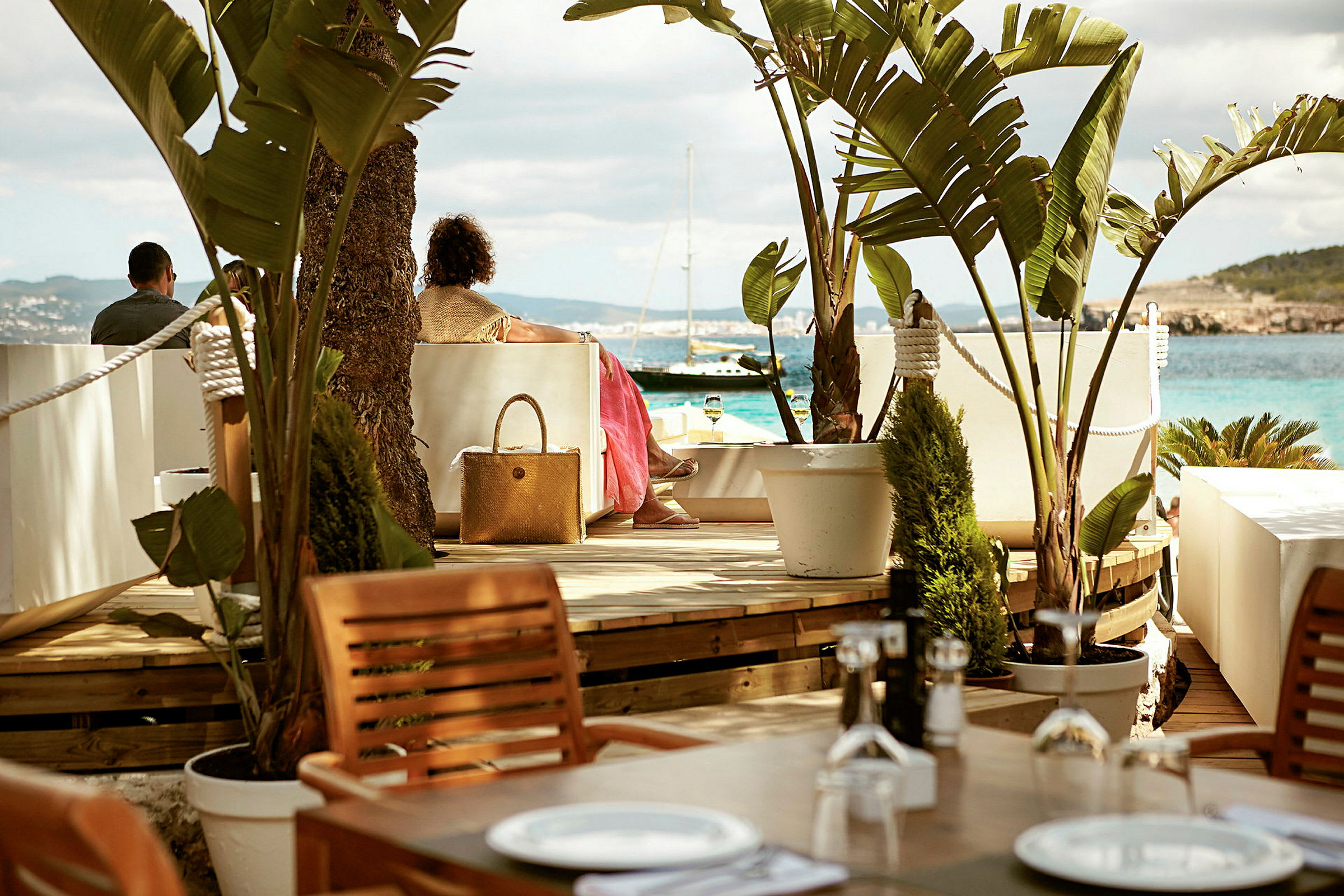 En udendørs terrasse med borde og stole omgivet af planter, hvor folk sidder og kigger ud over havet med en sejlbåd. (AI-genereret tekst)