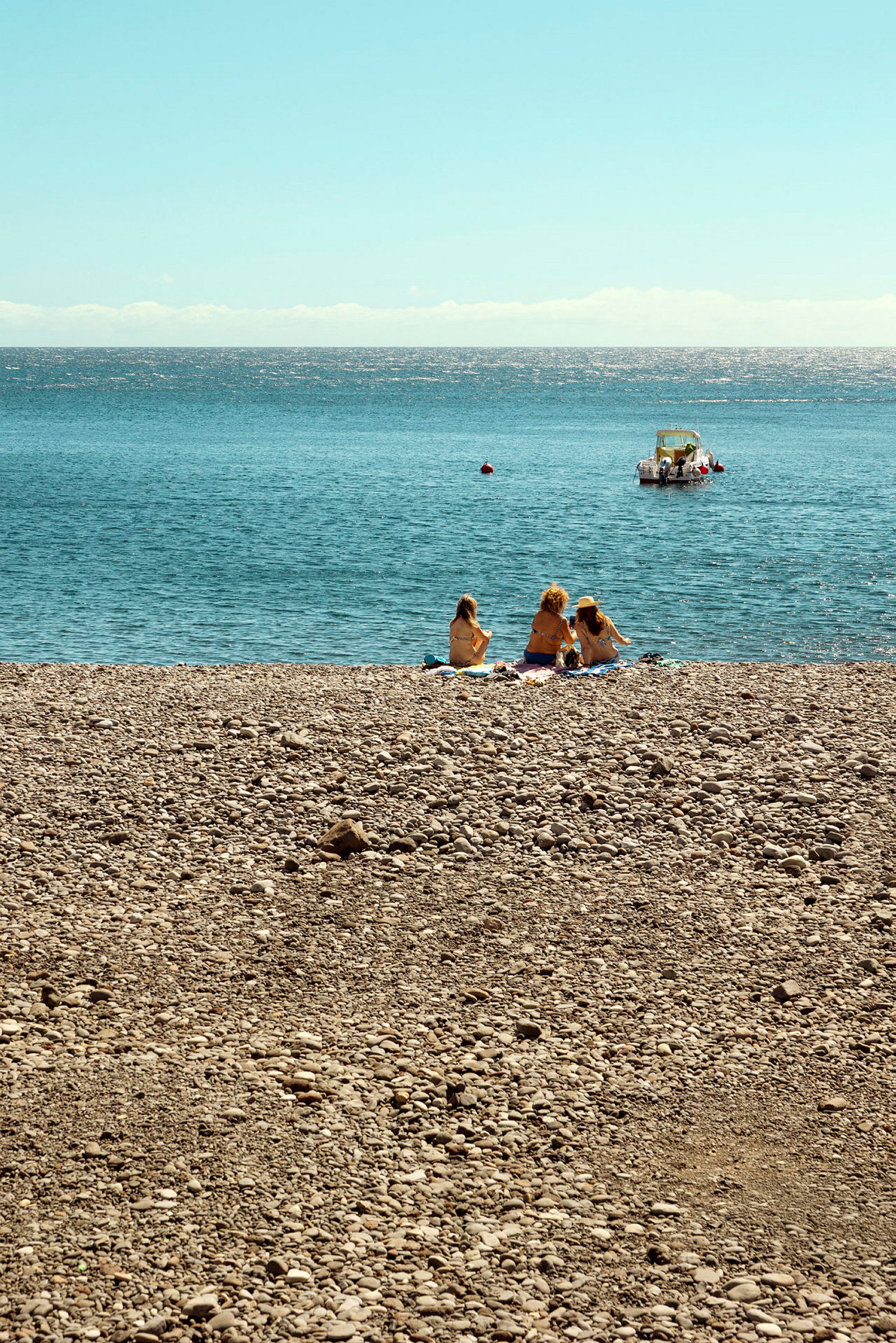 Tre personer sidder på en stenstrand med havet i baggrunden og en båd længere ude på vandet. (AI-genereret tekst)