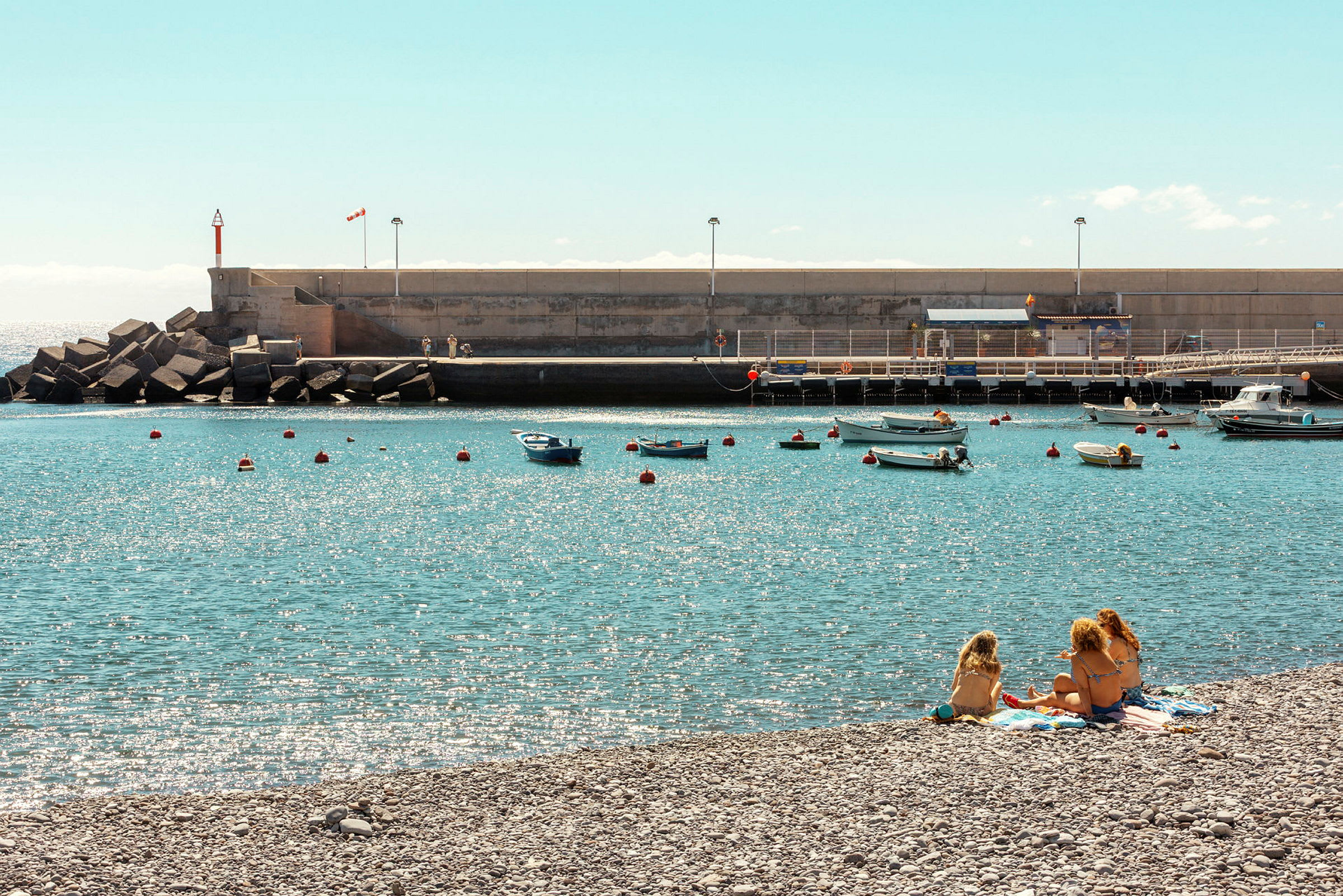 En strand med personer, der sidder på håndklæder ved vandet, med små både fortøjet i havnen. (AI-genereret tekst)