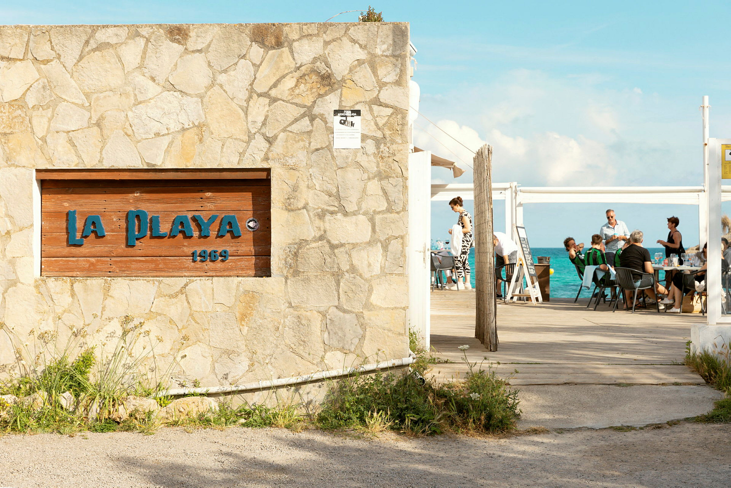 En stenmur med et skilt med teksten 'La Playa 1969' foran en udendørs servering med havet i baggrunden. (AI-genereret tekst)