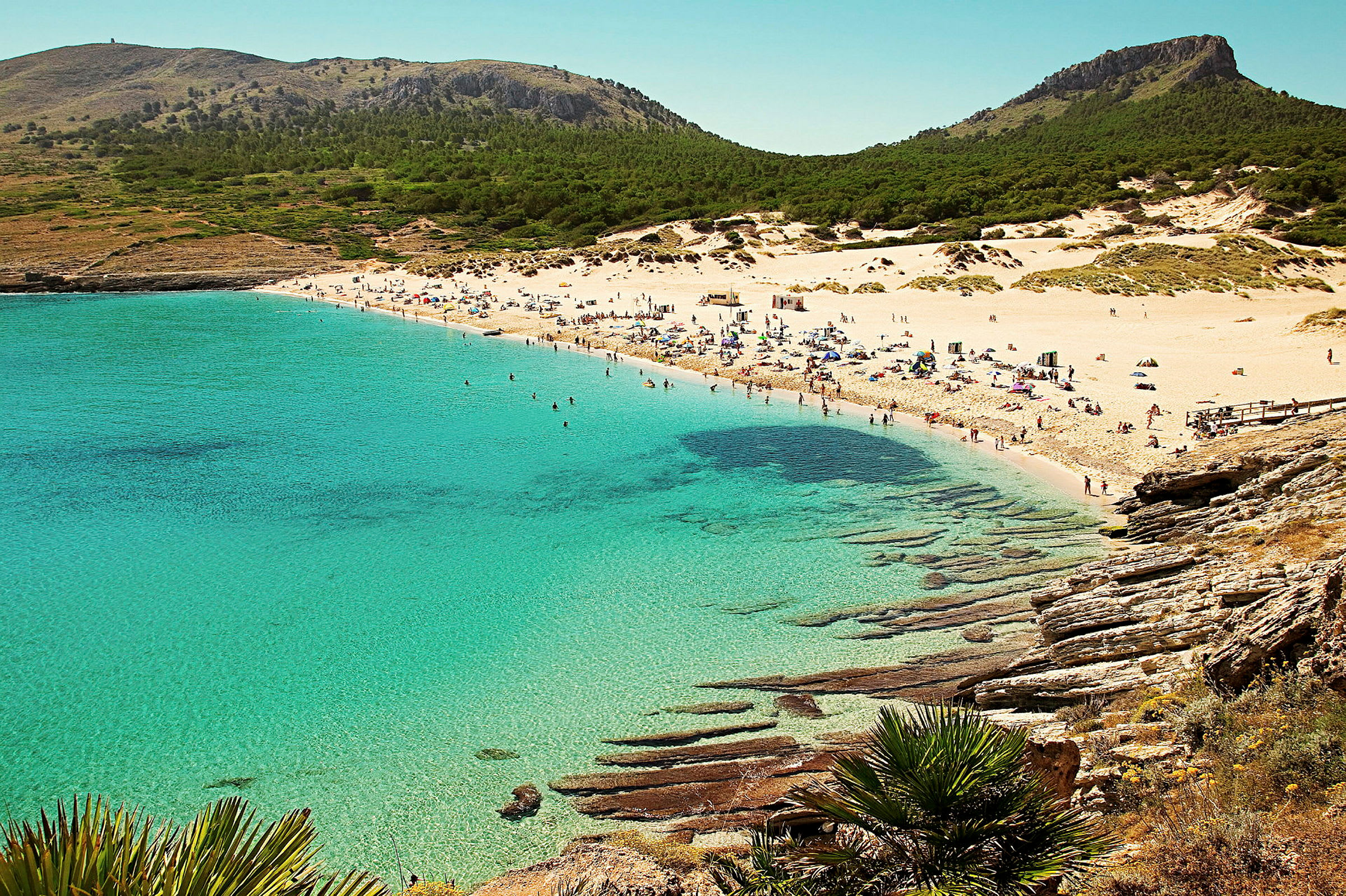 En sandstrand med krystalklart vand og mennesker, der soler sig og bader. Skovklædte bjerge ses i baggrunden. (AI-genereret tekst)