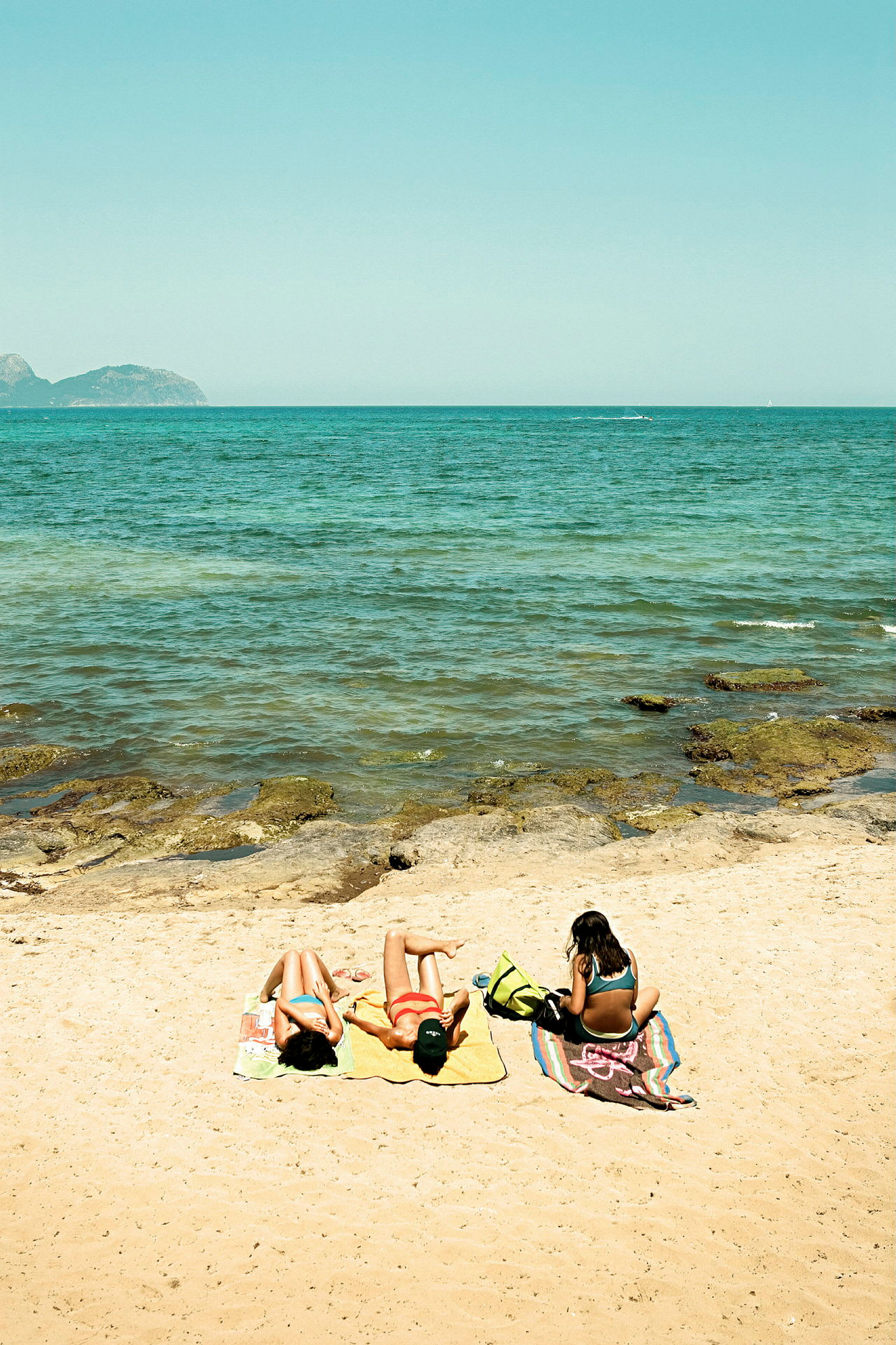 Tre personer ligger på håndklæder ved en sandstrand ved siden af havet. (AI-genereret tekst)
