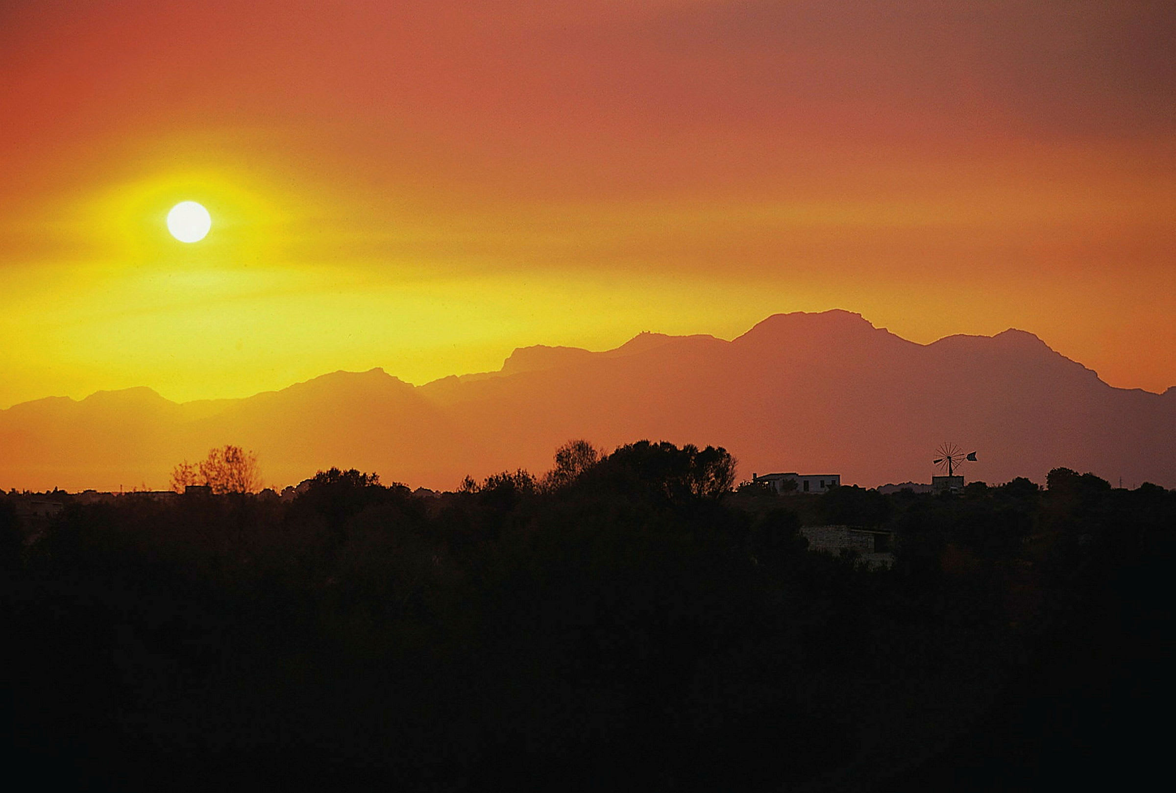 Solnedgang over silhuetten af et bjerg med træer og bygninger i forgrunden. (AI-genereret tekst)