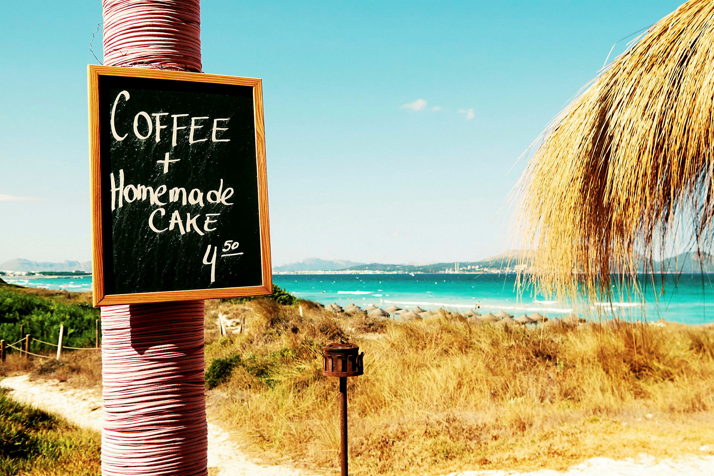 Skilt med teksten 'Coffee + Homemade Cake 4.50' ved stranden. (AI-genereret tekst)