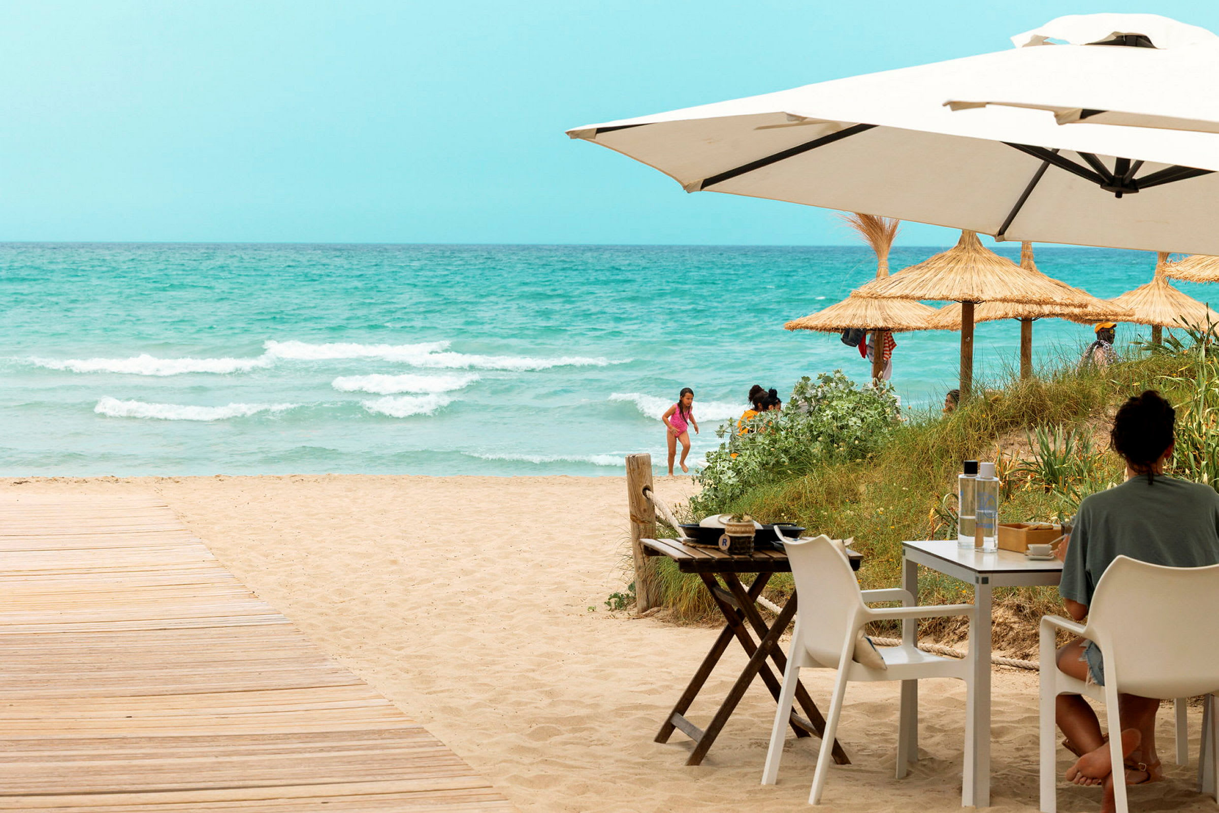 En person sidder ved et bord under en parasol ved en sandstrand med havet og strandparasoller i baggrunden. (AI-genereret tekst)