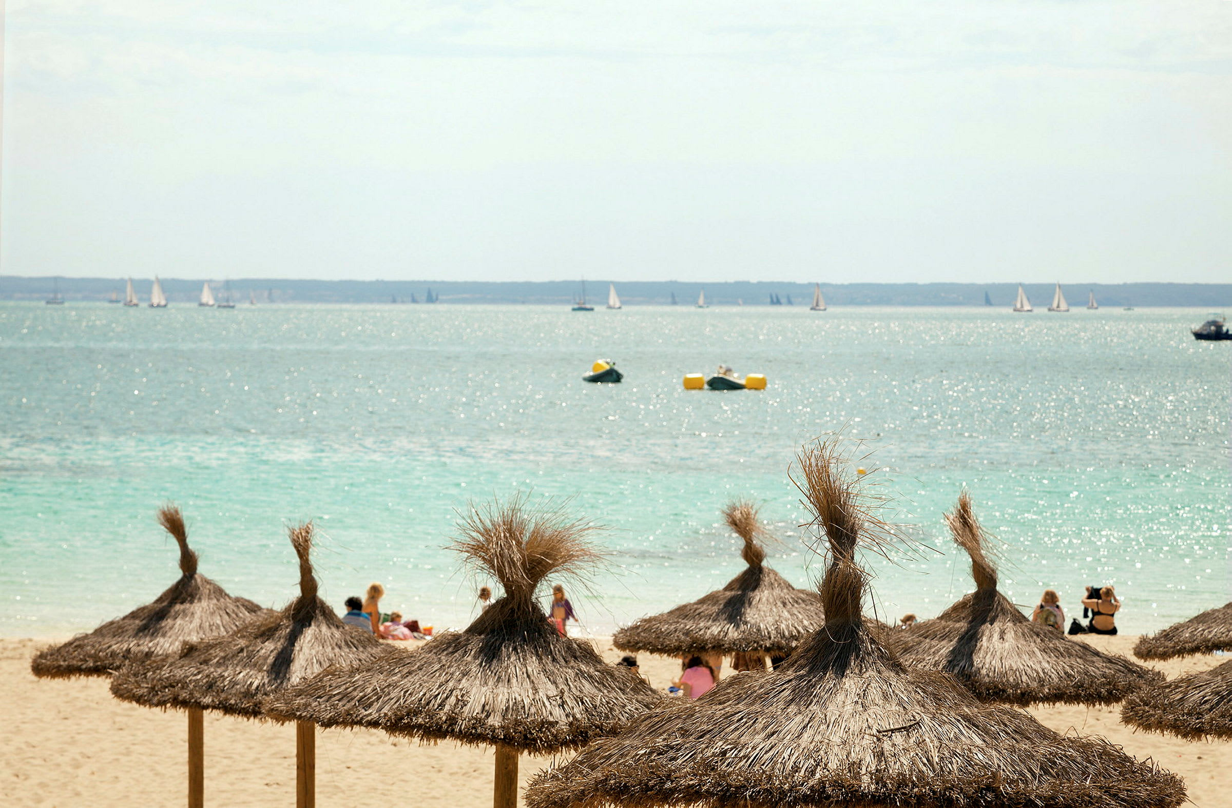 En strand med parasoller af tør halm, personer på stranden og en havudsigt med sejlbåde og flydende bøjer. (AI-genereret tekst)