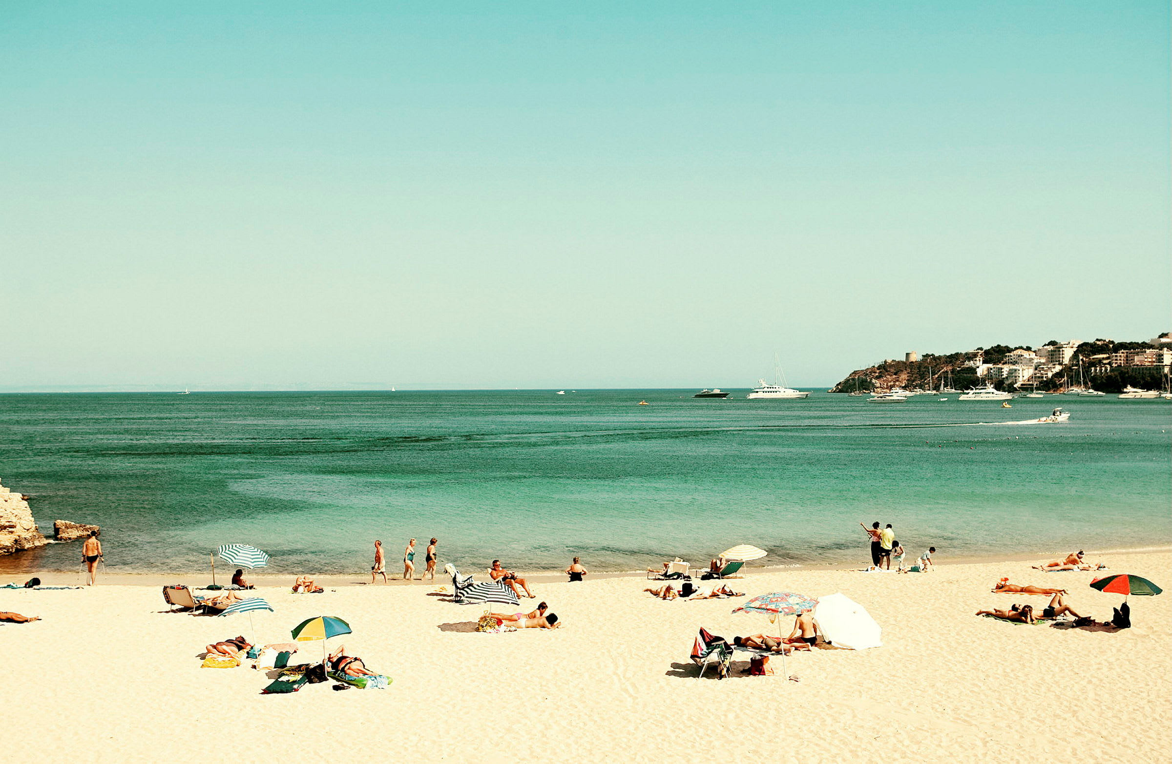 En strand med mennesker, der soler sig og bader, parasoller og havet med både i baggrunden. (AI-genereret tekst)