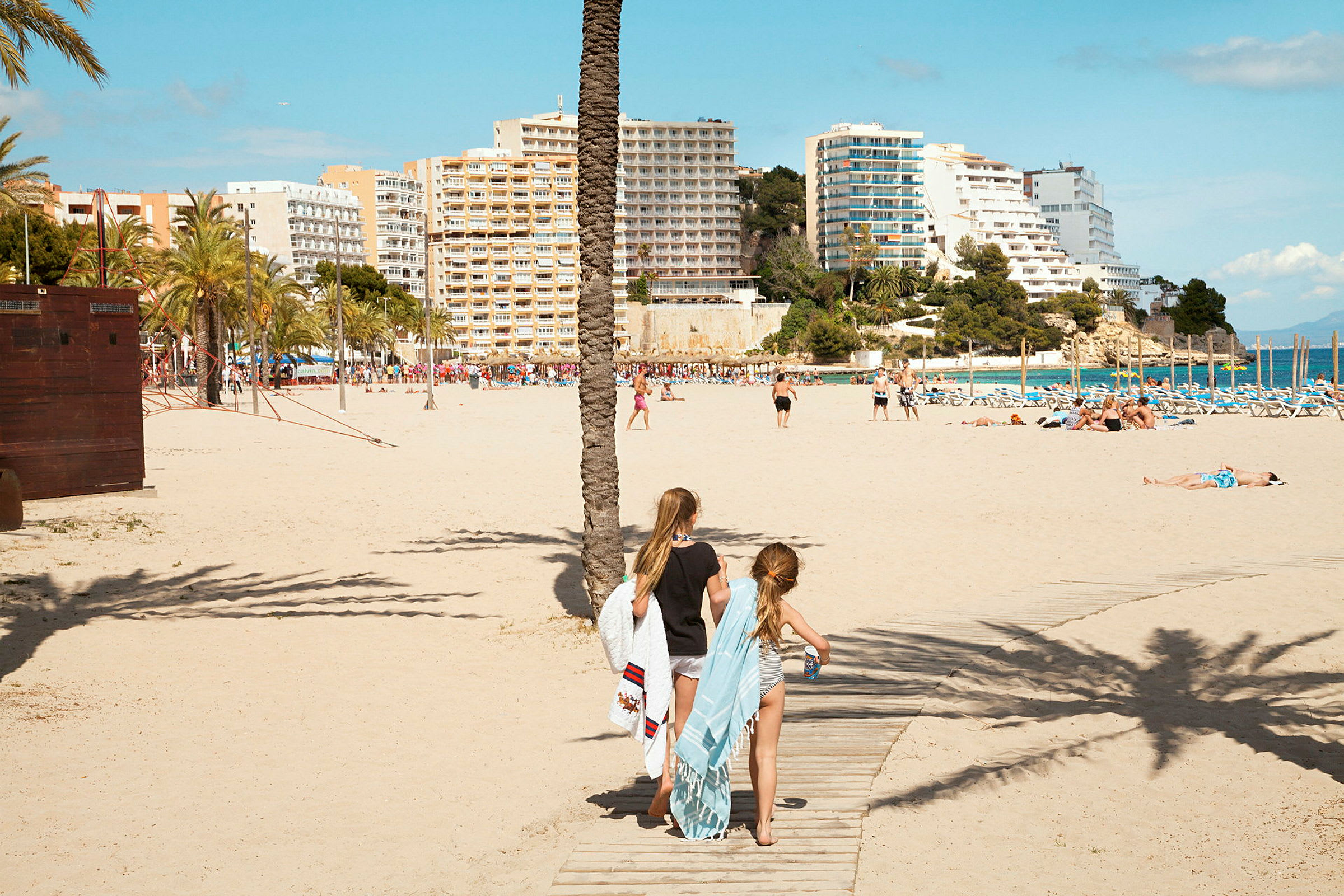 To personer går på en gangsti af træ på stranden, med håndklæder og badetøj. I baggrunden ses bygninger, palmer og havet. (AI-genereret tekst)