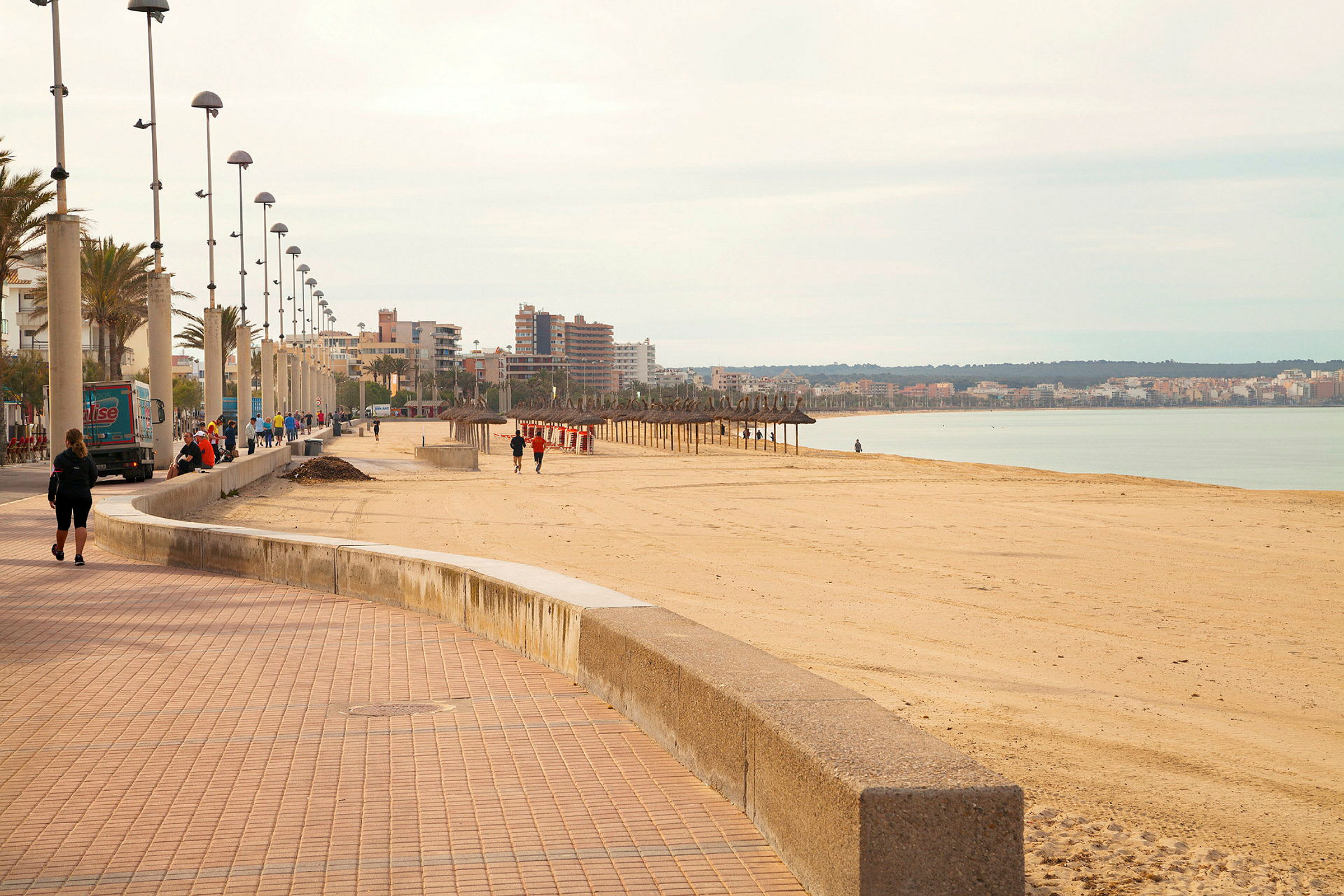 En bred strandpromenade med lygtepæle og mennesker, der går langs sandstranden. Byen og havet ses i baggrunden. (AI-genereret tekst)