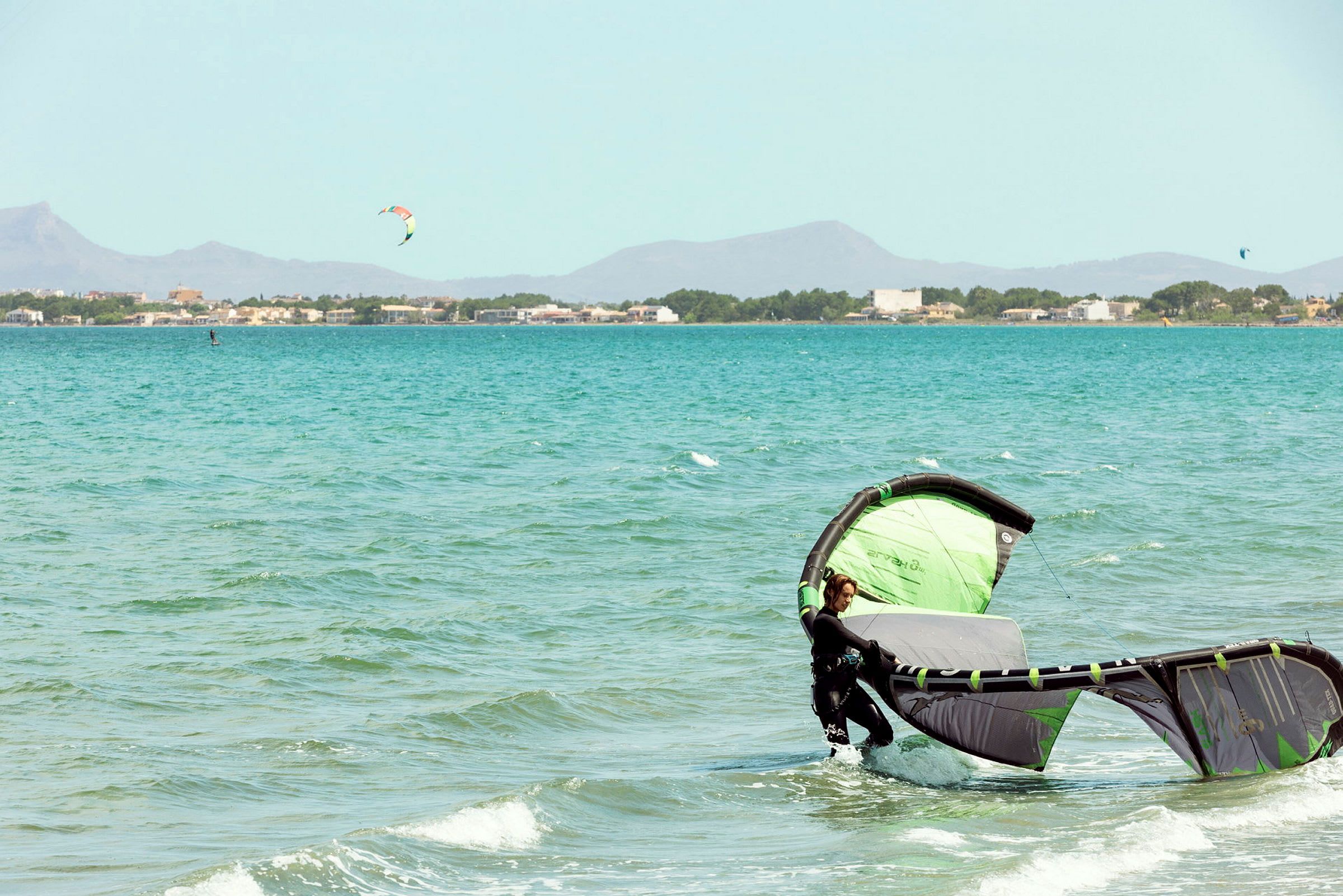 En person i vandet med en grøn og sort kitesurfing drage, med en by og bjerge i baggrunden. (AI-genereret tekst)