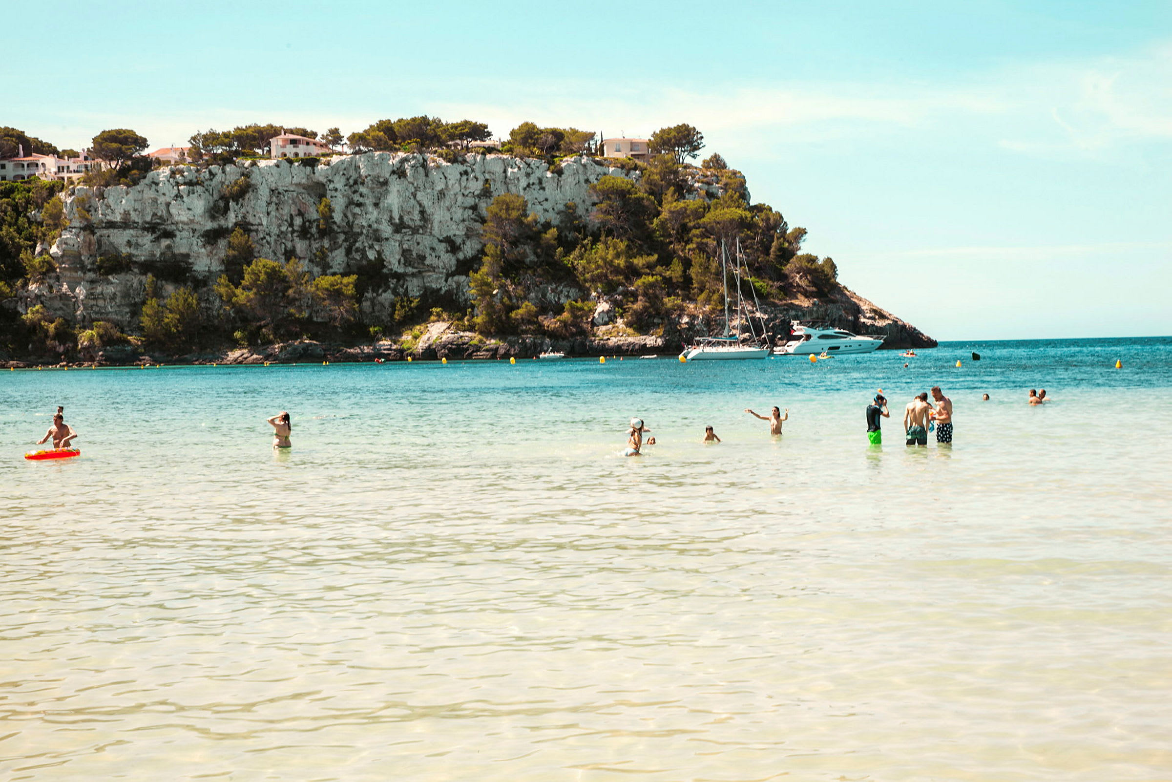 En strand med mennesker i vandet foran en bakke med klipper og nogle huse samt både i baggrunden. (AI-genereret tekst)