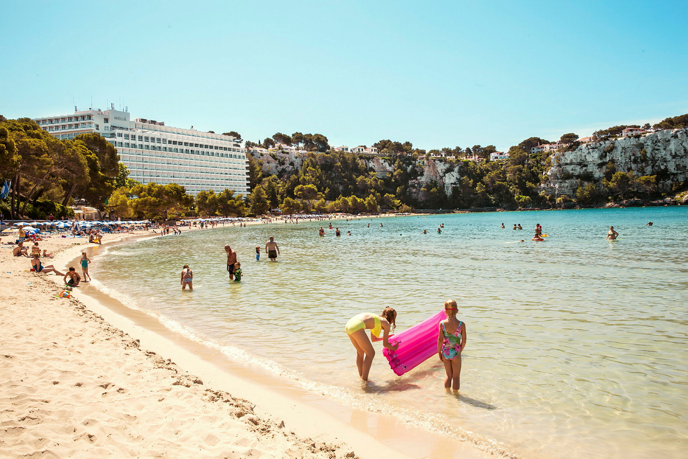 Strand med mennesker, der bader og leger i vandet foran en naturskøn klippe og et hotel. (AI-genereret tekst)