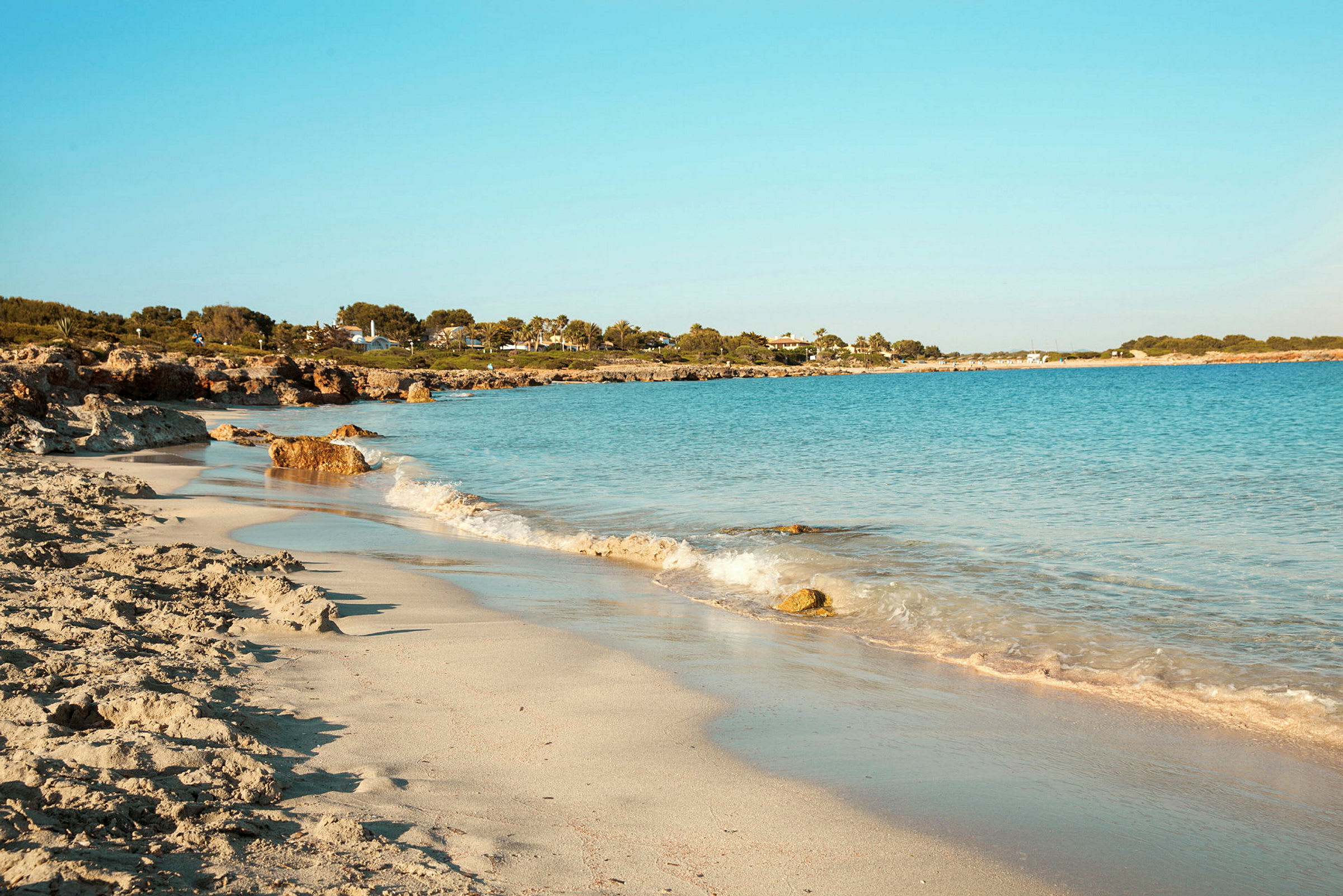 En strand med lys sand og blåt hav, omgivet af grønt og bygninger i baggrunden. (AI-genereret tekst)
