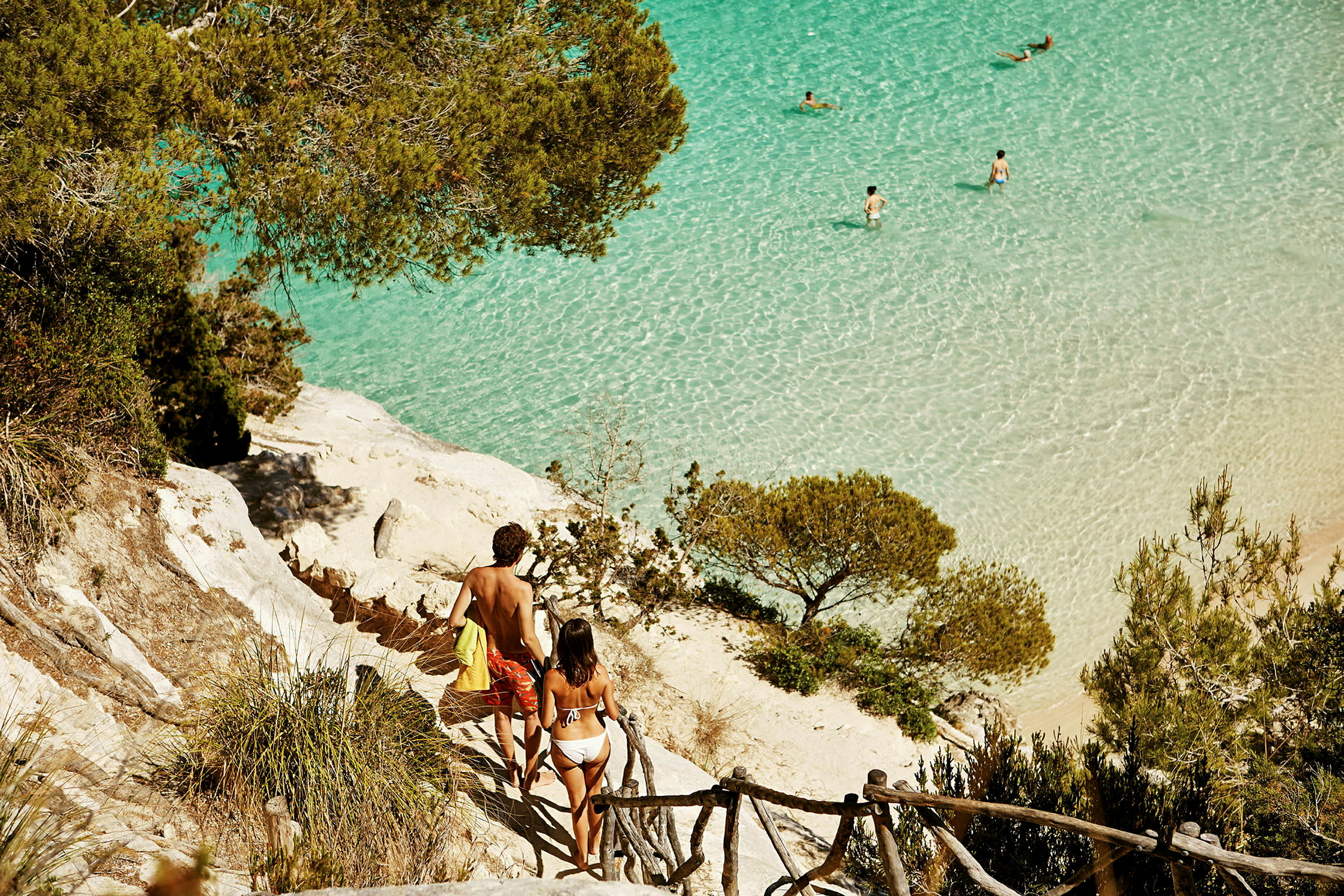 To personer går ned ad en stensti mod en strand med klart turkis vand, omgivet af vegetation. (AI-genereret tekst)