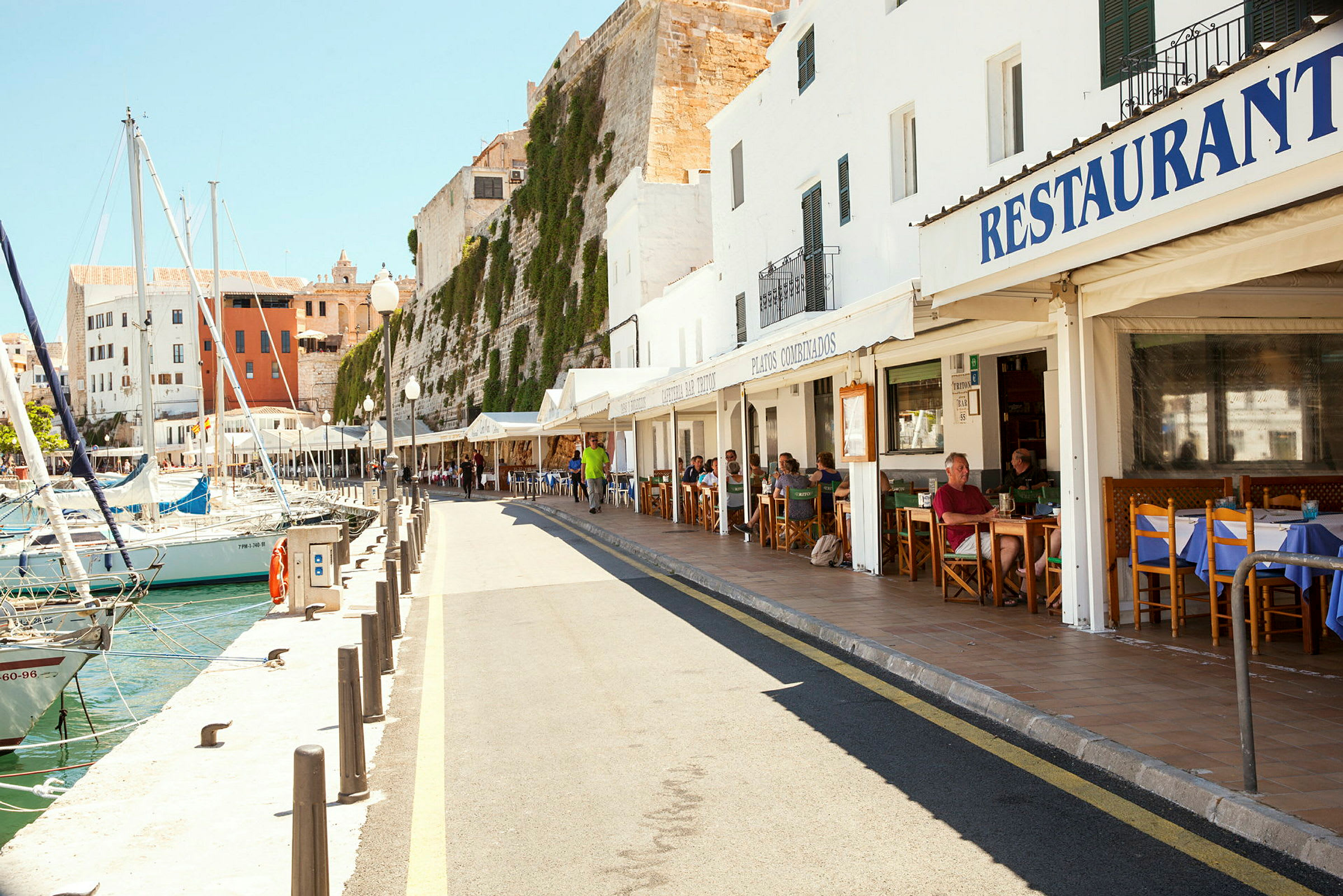 En havnepromenade med restauranter og både ved kajen. Det er en solrig dag med mennesker, der sidder udenfor på restauranternes terrasser. (AI-genereret tekst)