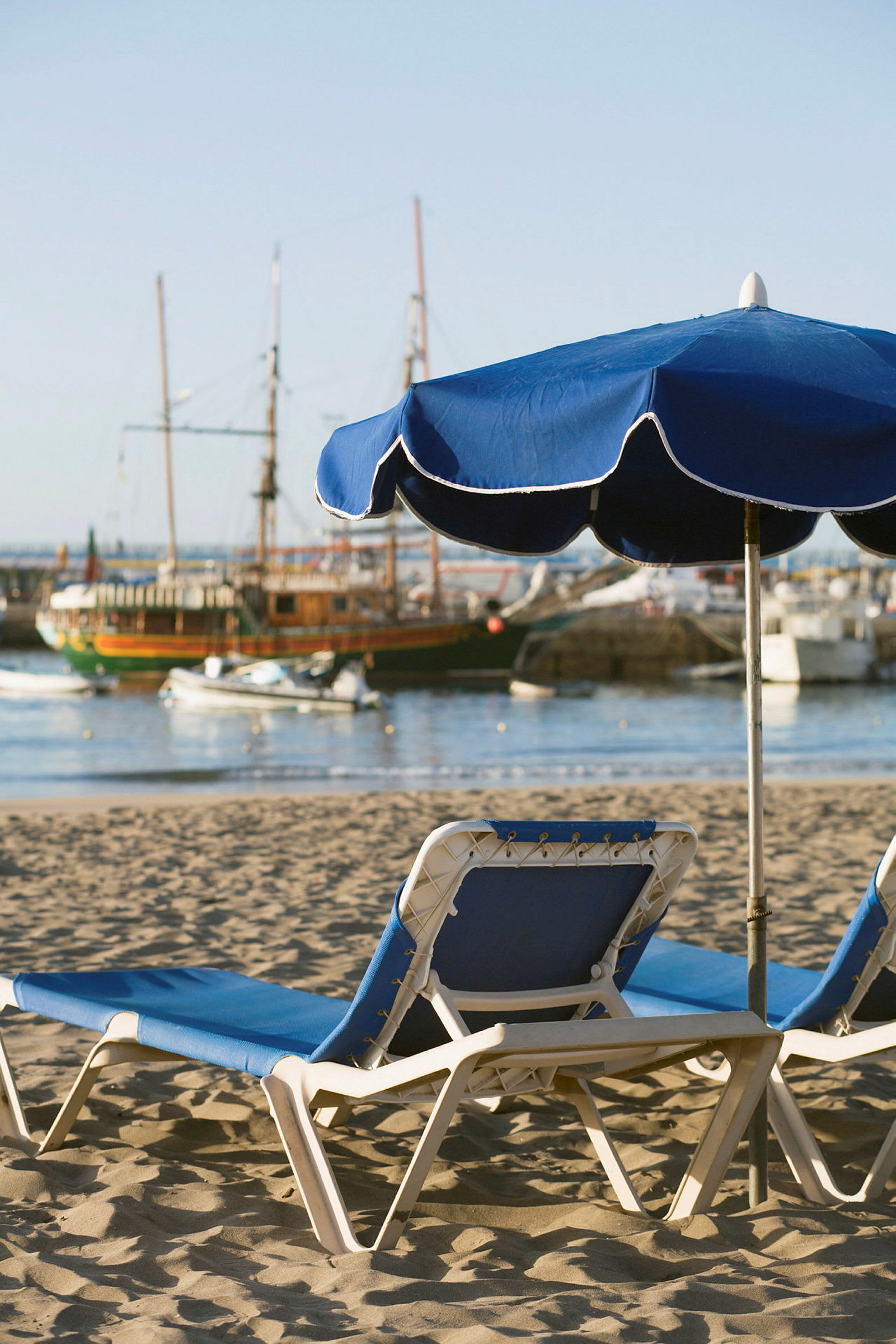 To solvogne med blå puder og en blå parasol på en sandstrand med sejlbåde i baggrunden. (AI-genereret tekst)