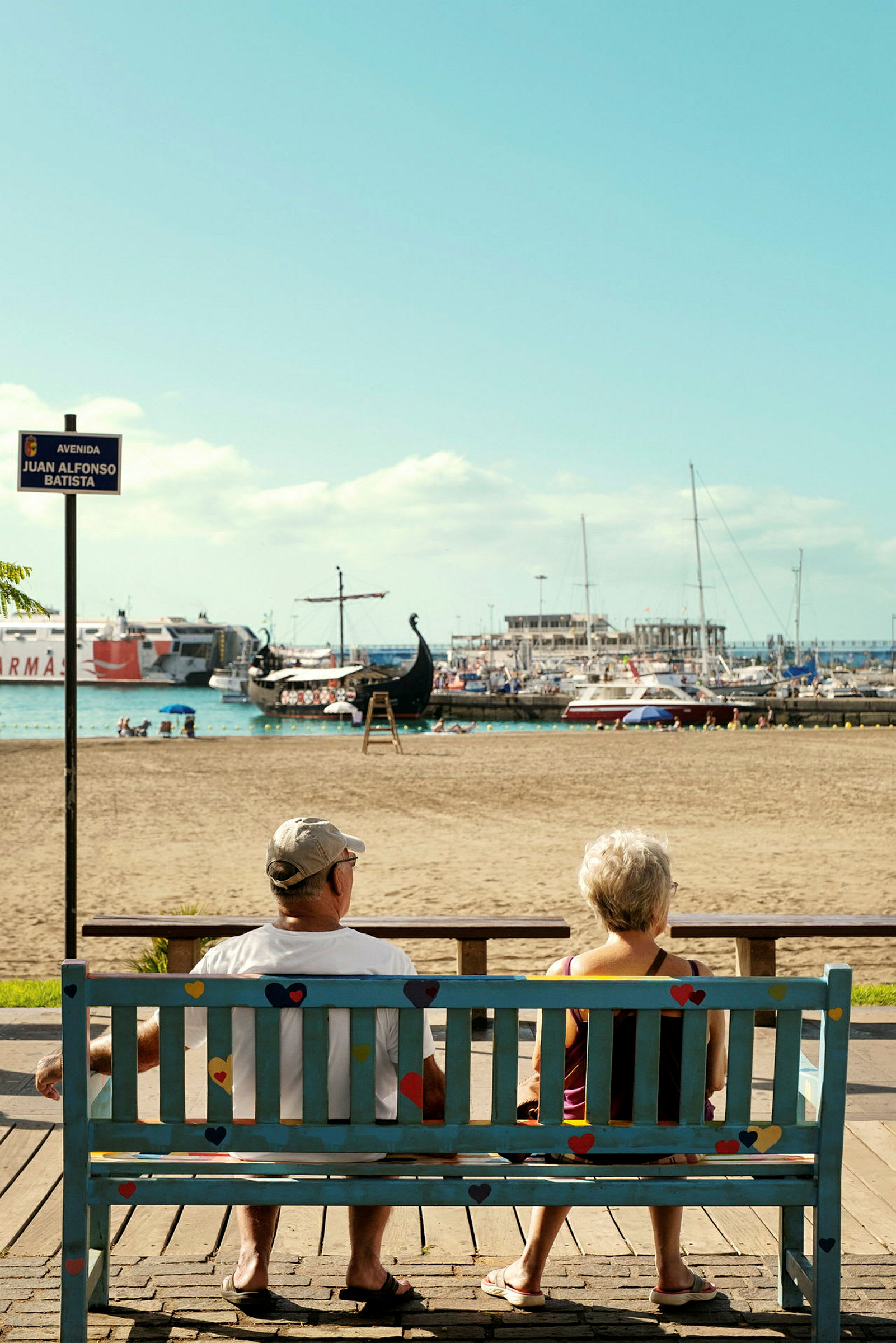 To personer sidder på en bænk med udsigt over stranden og havnen. (AI-genereret tekst)