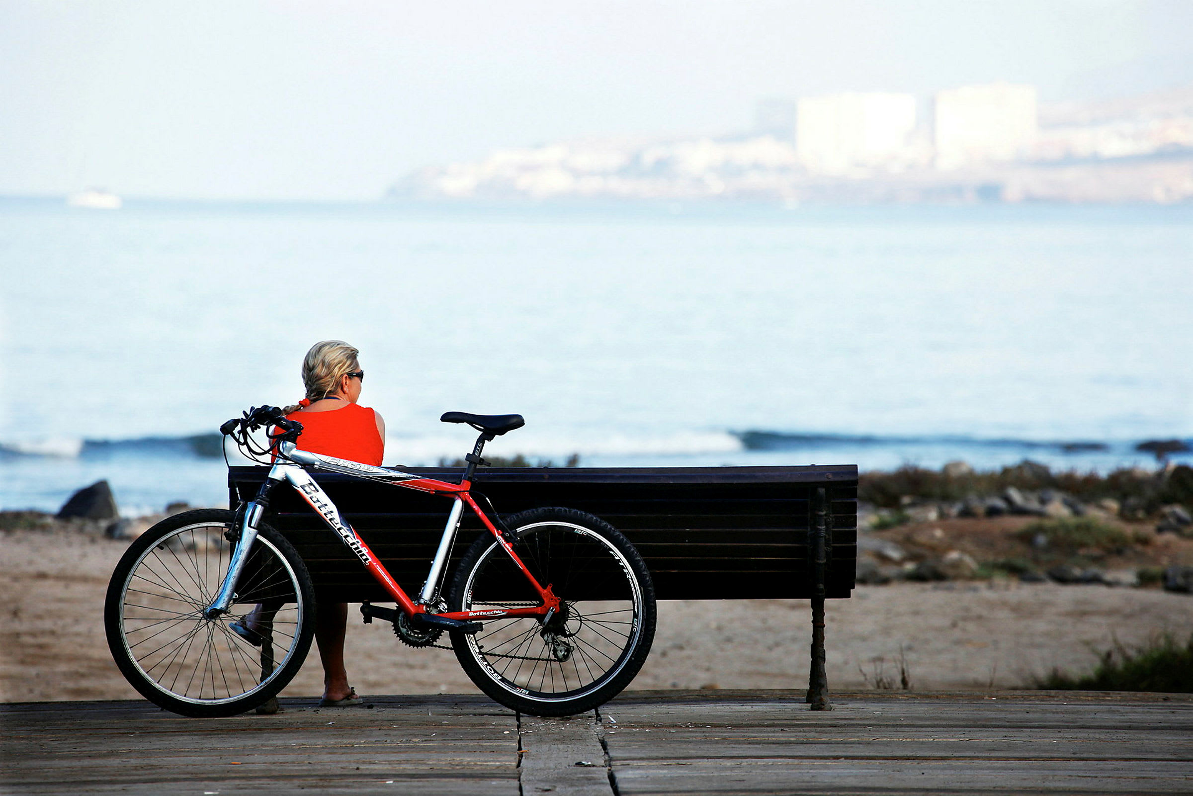 En person sidder på en bænk ved stranden med en cykel ved siden af. (AI-genereret tekst)