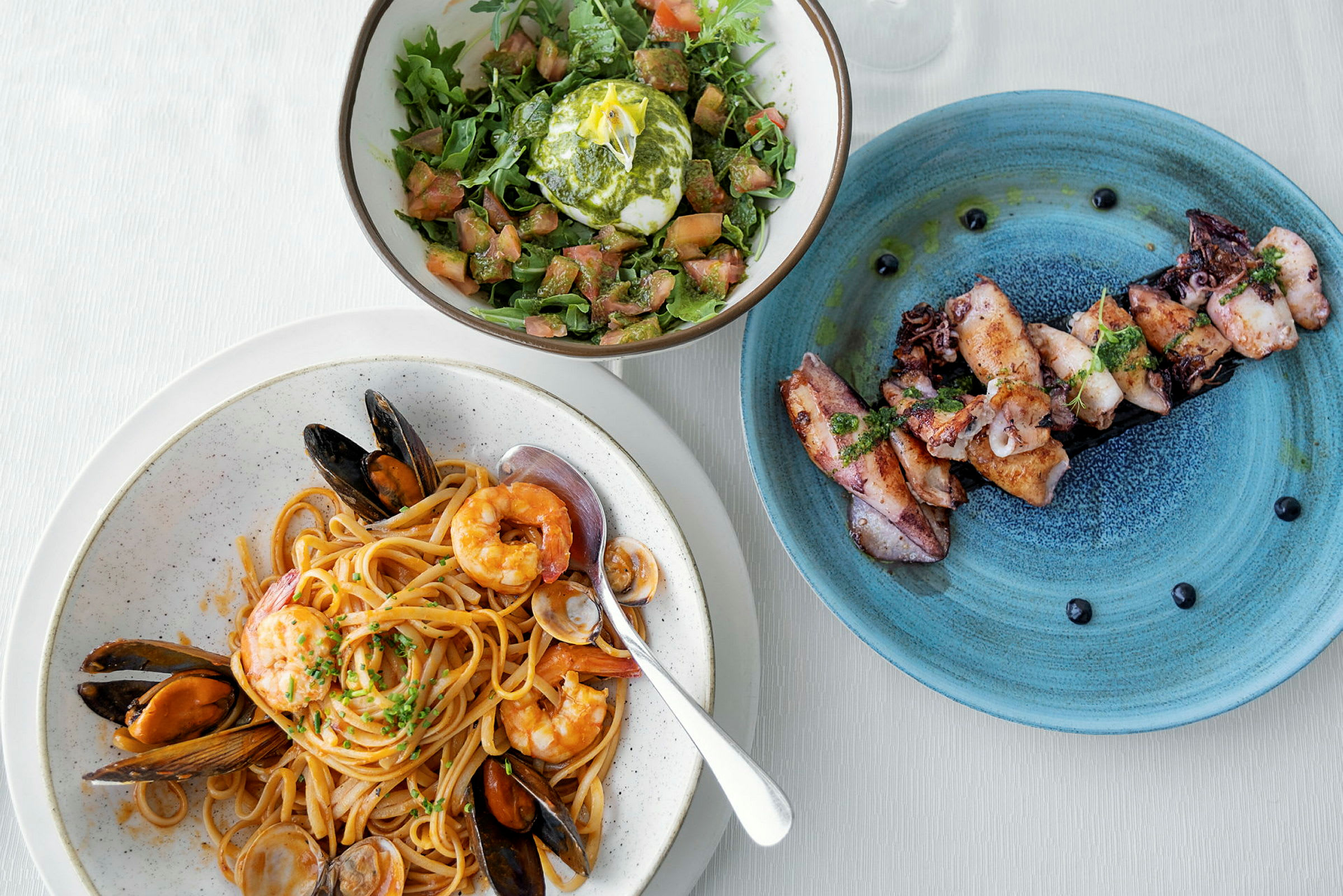 En tallerken med pasta og skaldyr, en salat med mozzarella og en tallerken med grillet blæksprutte. (AI-genereret tekst)