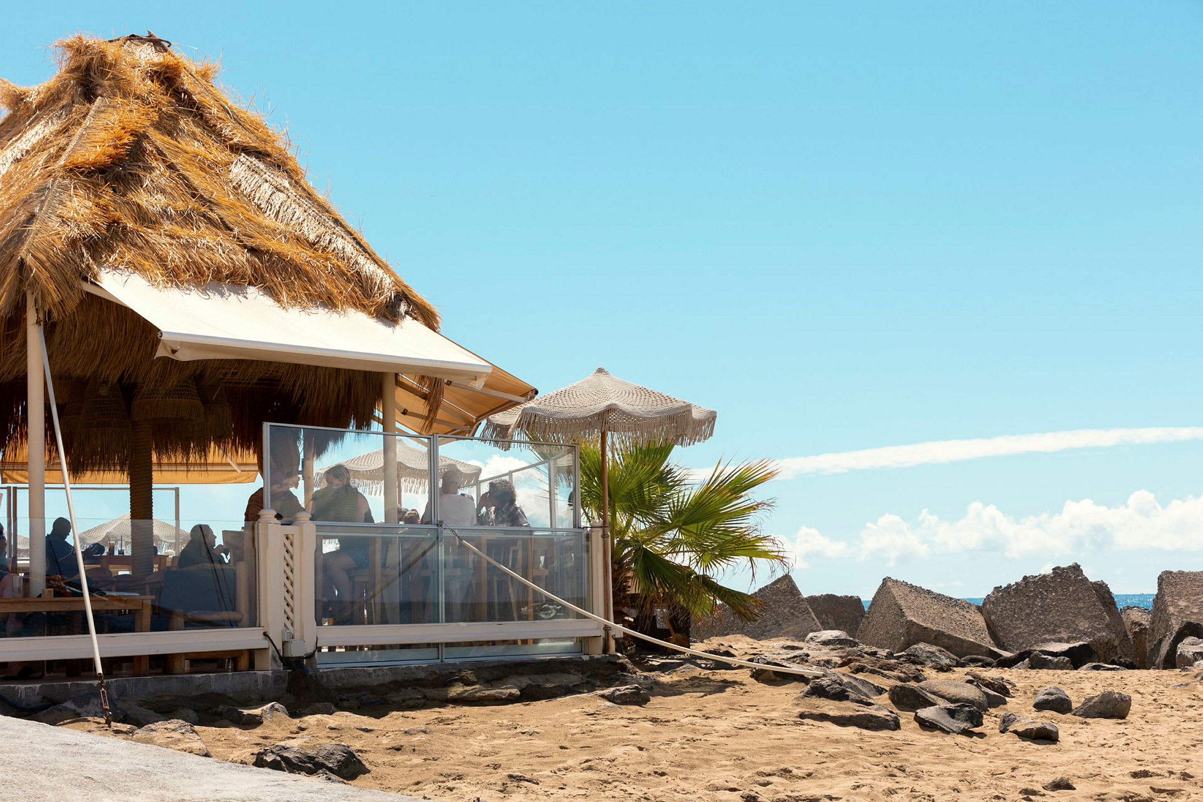 En strandbar med stråtag og parasoller, omgivet af sand og klipper. (AI-genereret tekst)