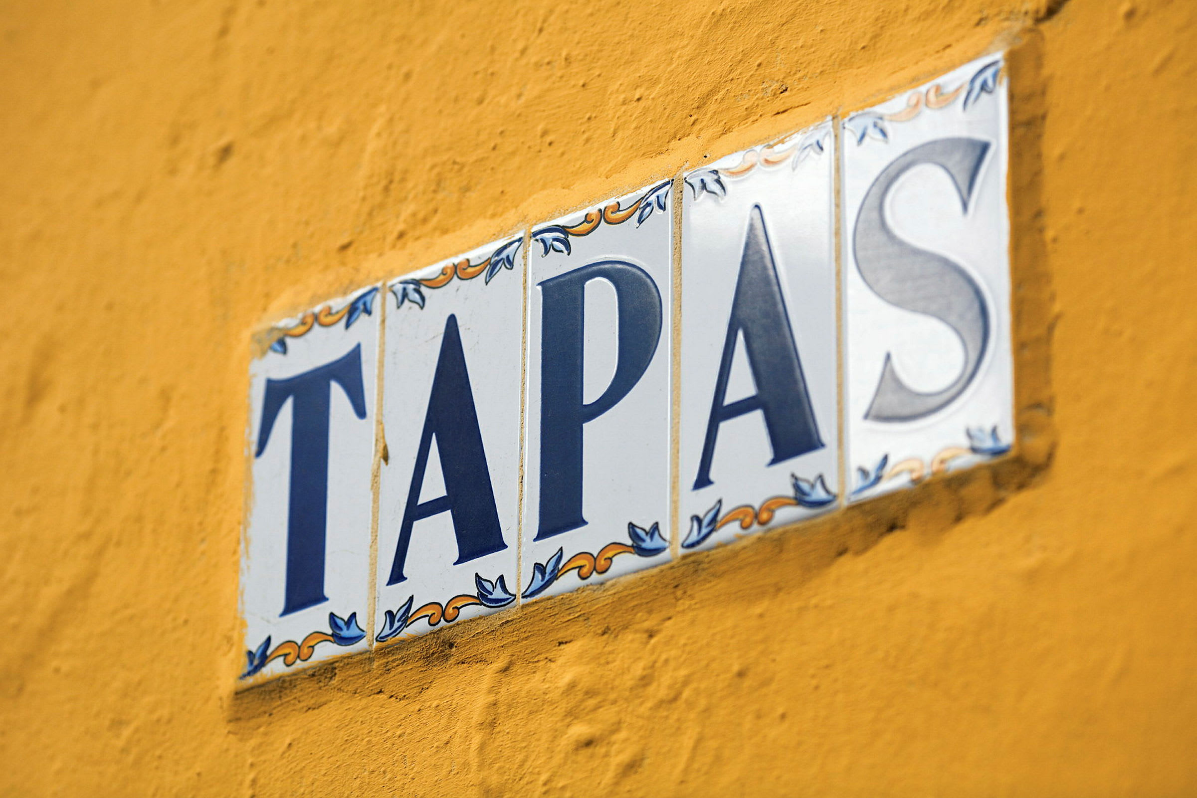 Et skilt med teksten 'TAPAS' på hvide keramiske fliser mod en gul væg. (AI-genereret tekst)