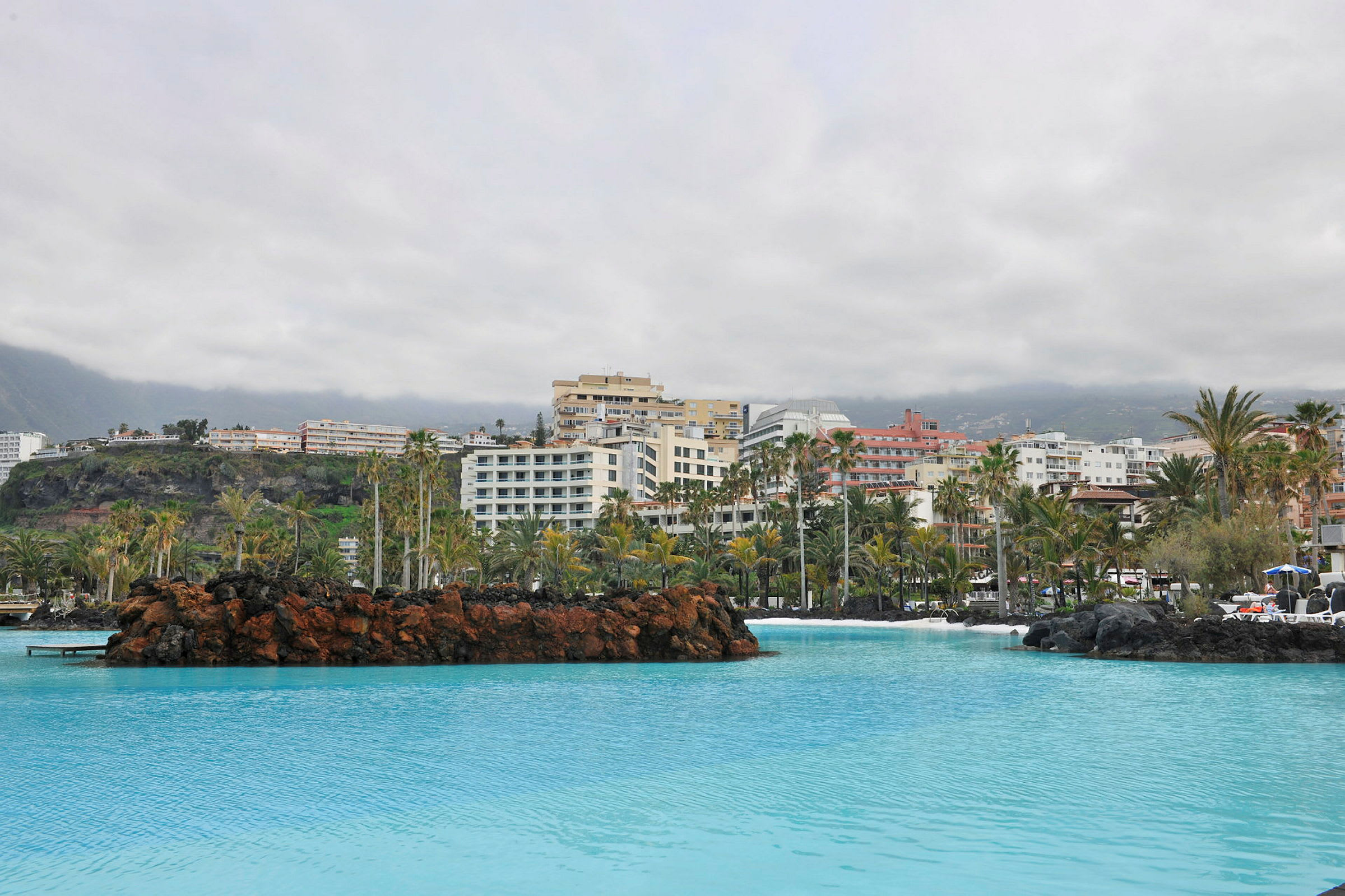 Hotelbygninger bag en pool med palmer og lavformationer. (AI-genereret tekst)