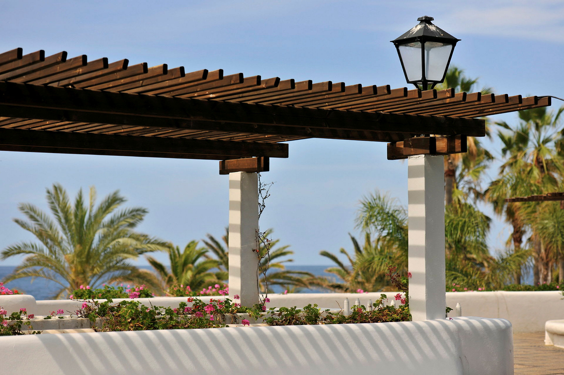 Pergola med træbjælker og blomsterdekorationer, palmer og hav i baggrunden. (AI-genereret tekst)