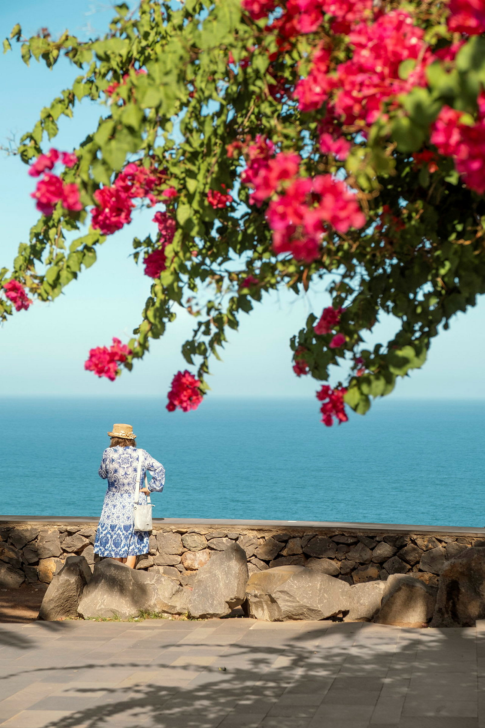 Person der står ved en stenmur med udsigt over havet under en blomstrende bougainvillea. (AI-genereret tekst)